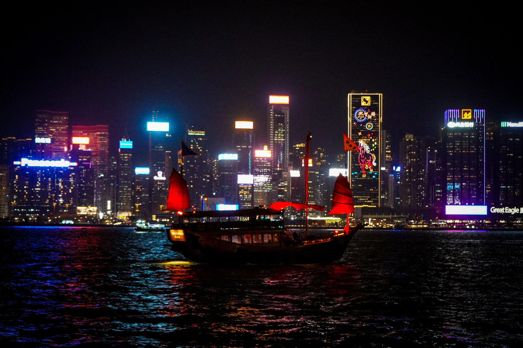 Night view of a city skyline with illuminated buildings and neon signs, reflecting on the water, with a traditional boat in the foreground.
