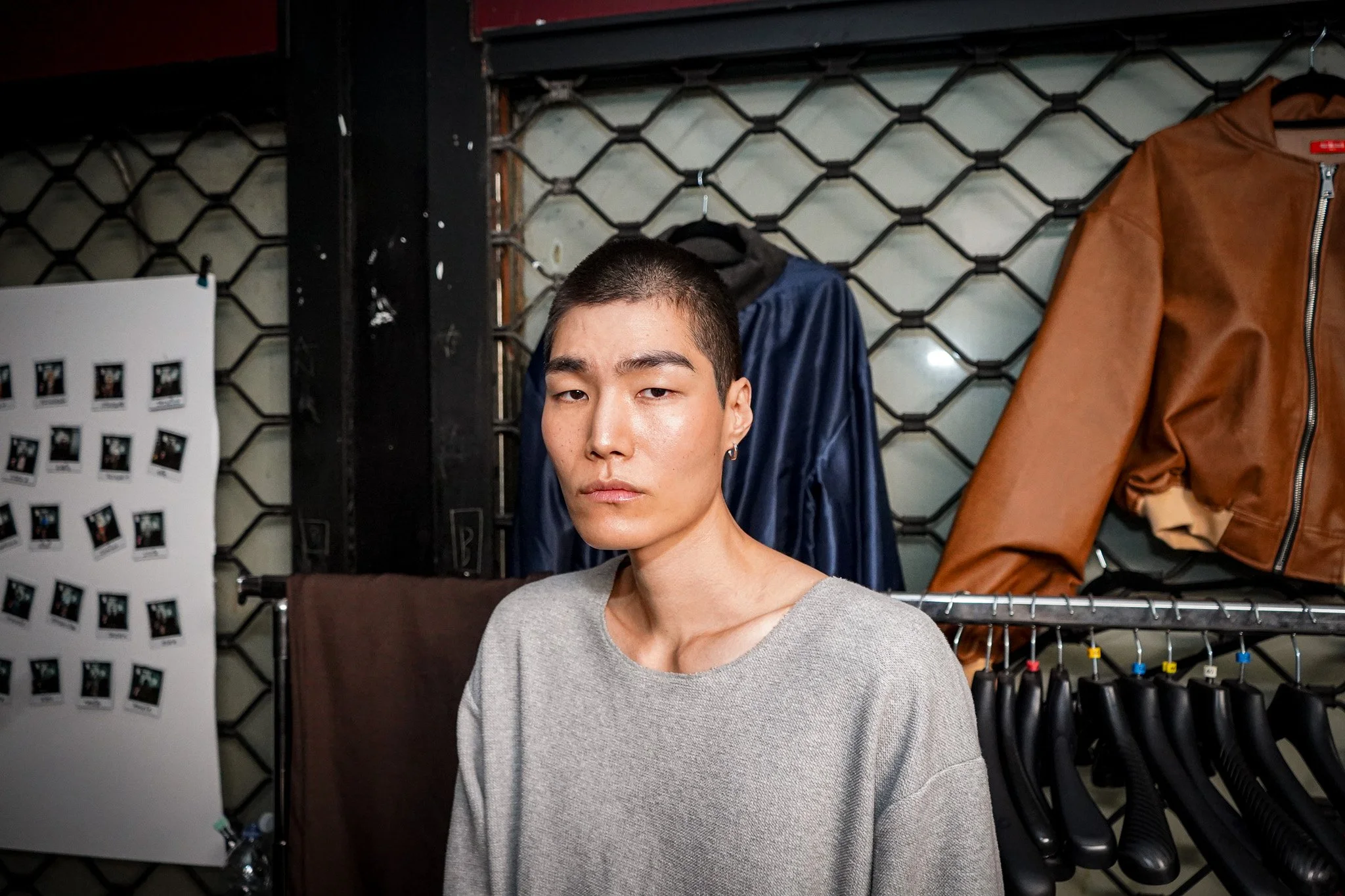 A young man with short black hair and a hoop earring in his left ear, wearing a light gray sweater, standing in front of a clothing rack with jackets and hanging items, indoor setting with a black grid wall and assorted photos nearby.