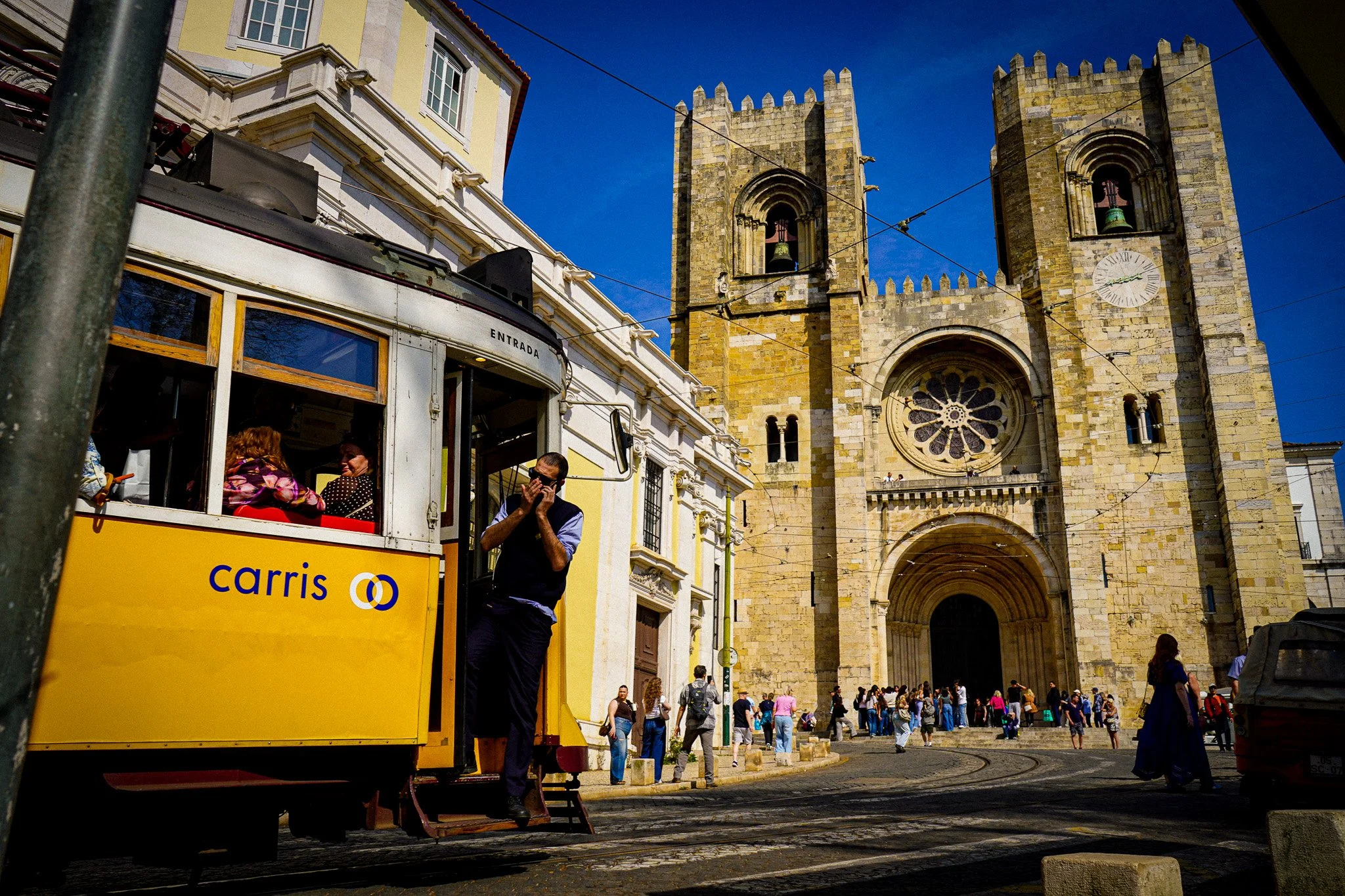 A yellow tram with the word 'carris' on it is stopped on a street in front of a historic stone church with two bell towers and an arched entrance. There are people walking and standing around, and a man is seen adjusting his face mask near the tram. 