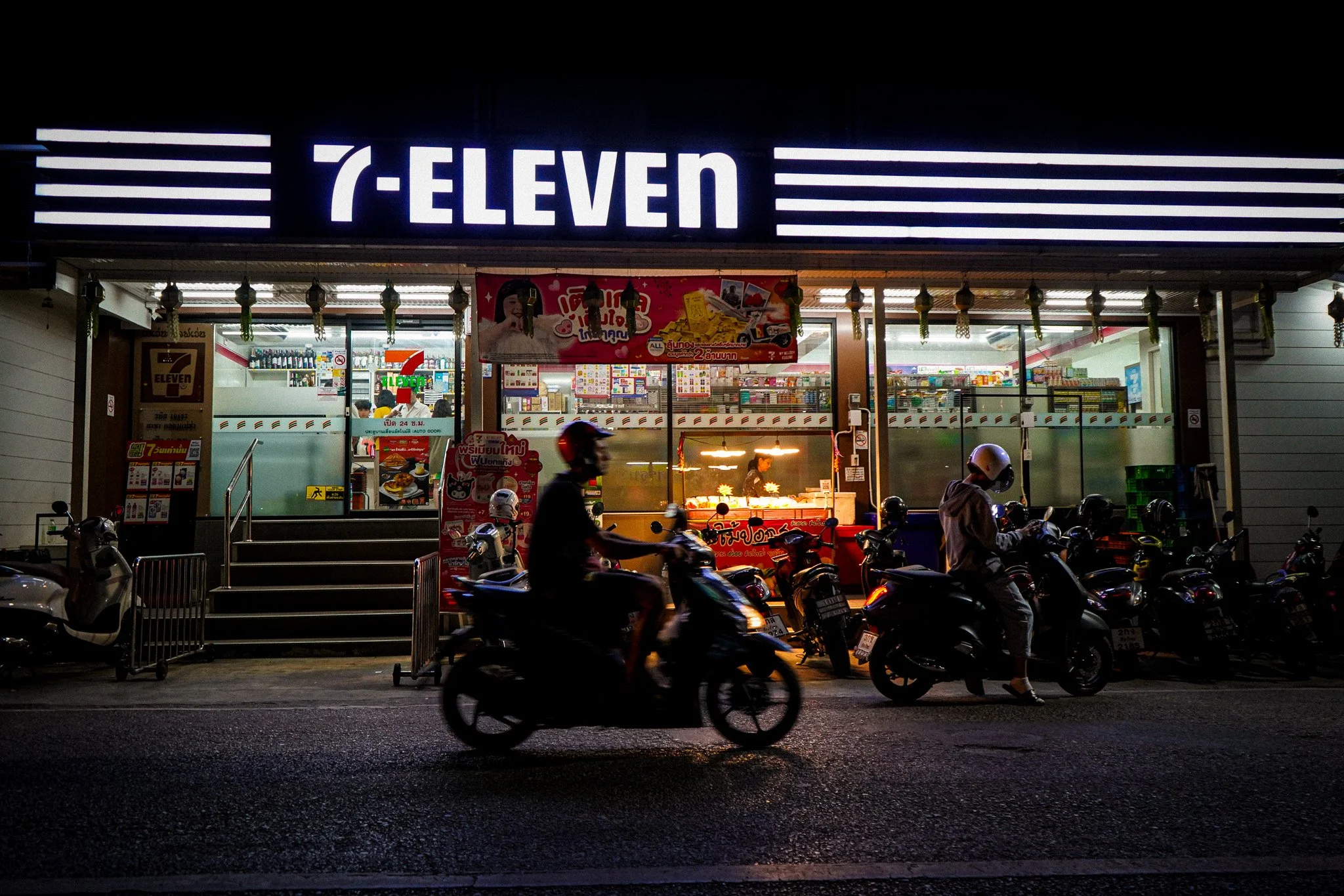 Night scene outside a 7-Eleven convenience store with people on motorbikes passing by on the street.