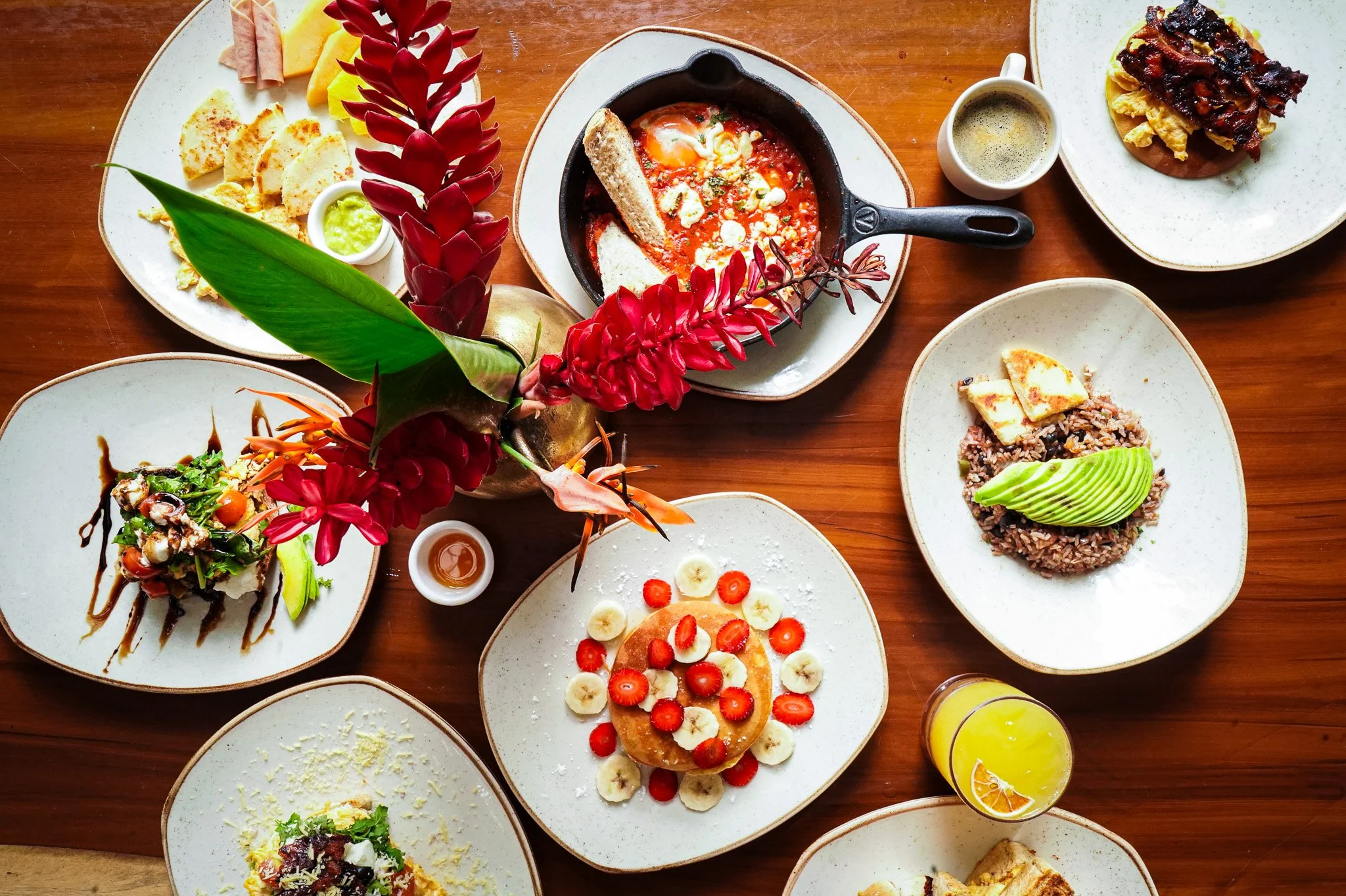 An overhead view of a variety of breakfast and brunch dishes on a wooden table, including a slice of French toast topped with strawberries and banana slices, a pancake with syrup, salads, a baked egg dish in a skillet, and beverages, with red floral 