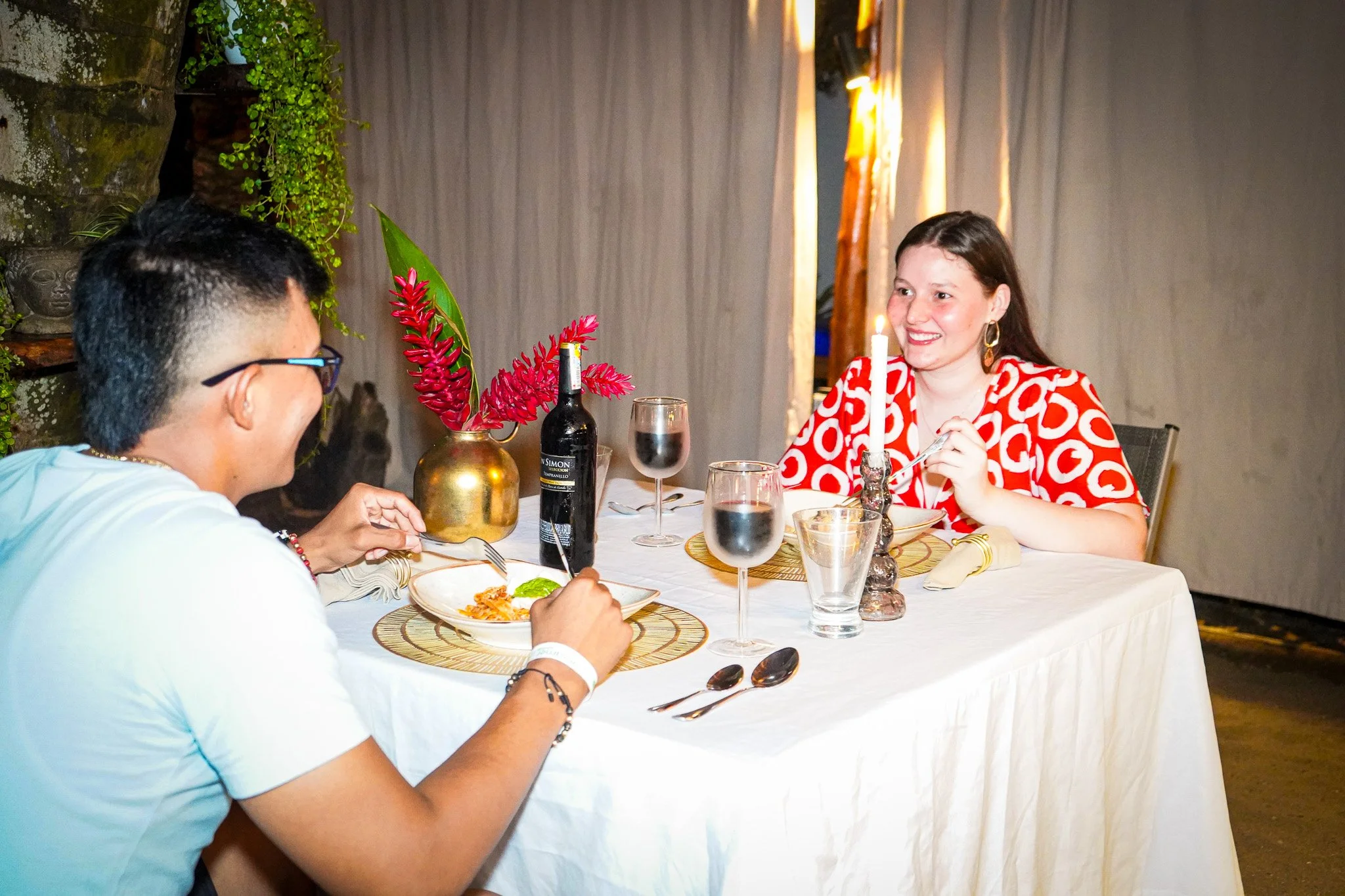 A woman with long brown hair and earrings is smiling while sitting at a dinner table with a man wearing glasses. The table has a white tablecloth, glasses of red wine, a plate of food, a decorative plant, and a lit candle. The background features bei