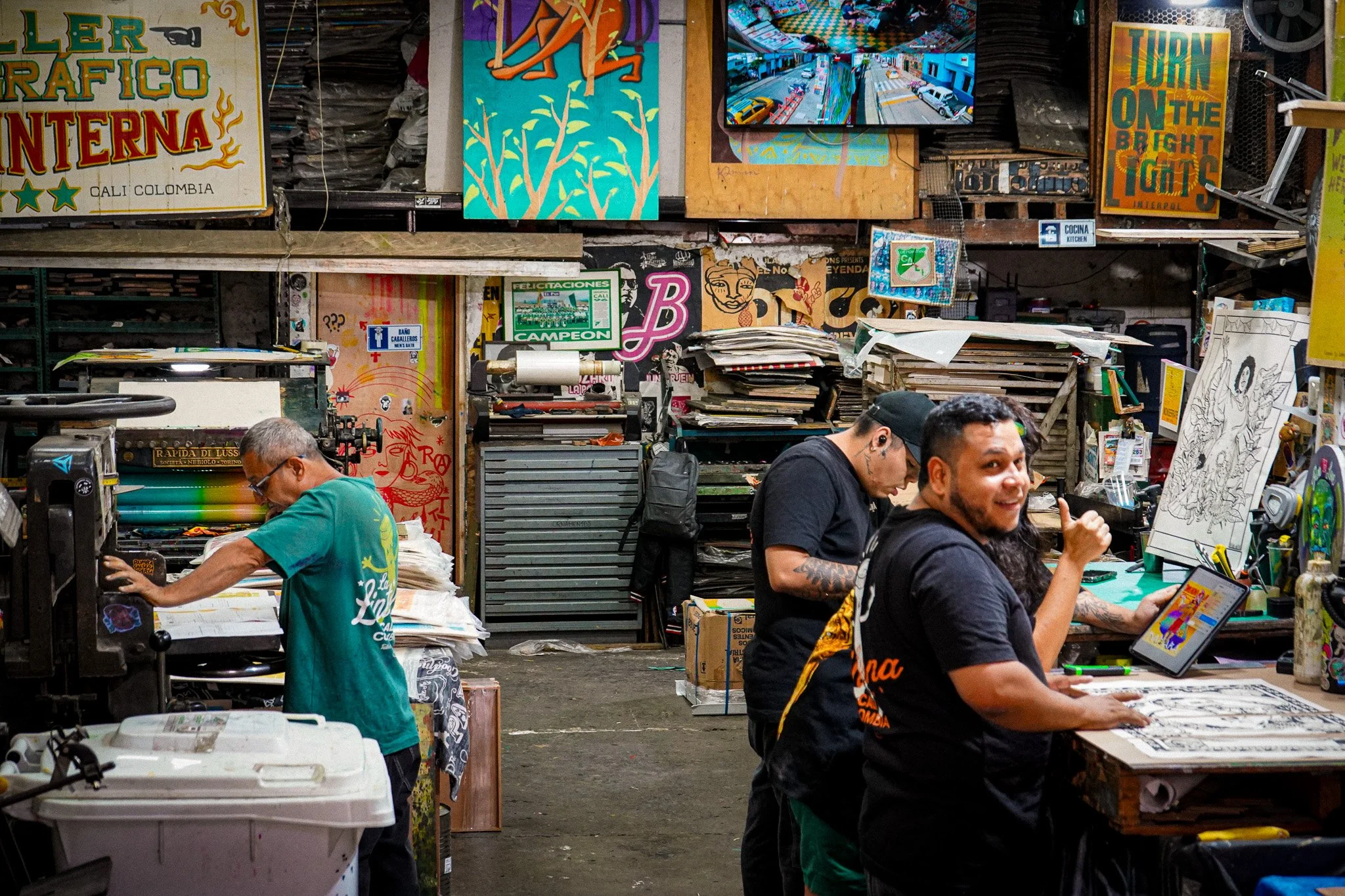 People working and socializing in an indoor art market or craft fair with colorful paintings, artwork, and signs on shelves and walls.