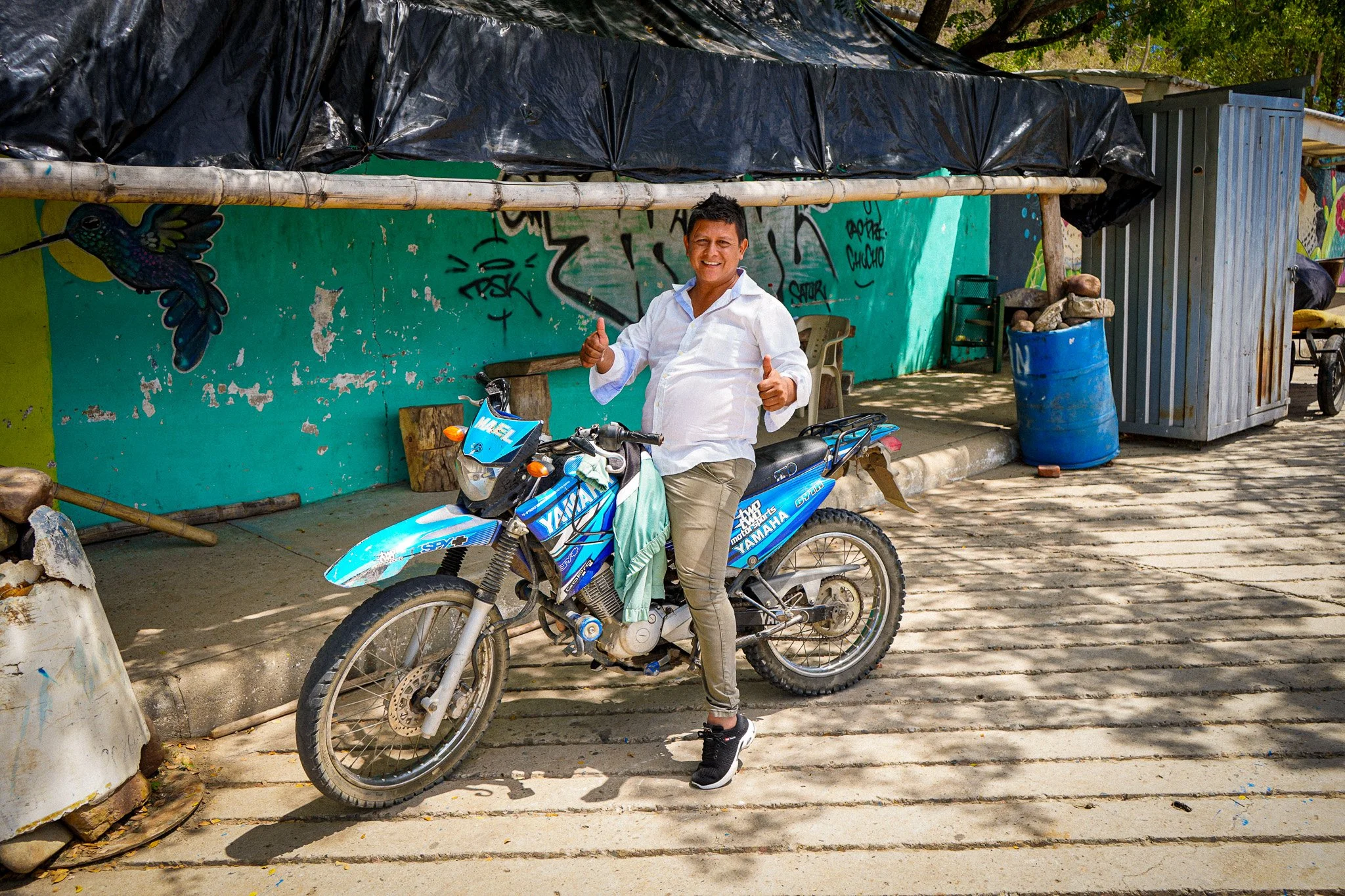 A man in a white button-up shirt, beige pants, and black sneakers is smiling and giving two thumbs up while standing next to a blue Yamaha dirt bike. Behind him is a colorful graffiti-covered wall, a plastic chair, a blue barrel, a small structure wi