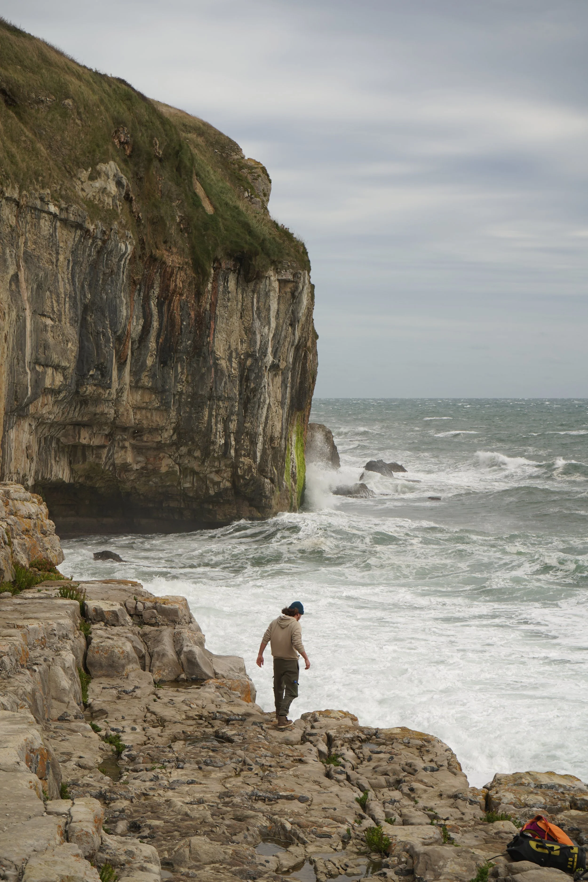 A person wearing a hoodie and hat walking on rocky shoreline beside ocean with high cliffs and overcast sky