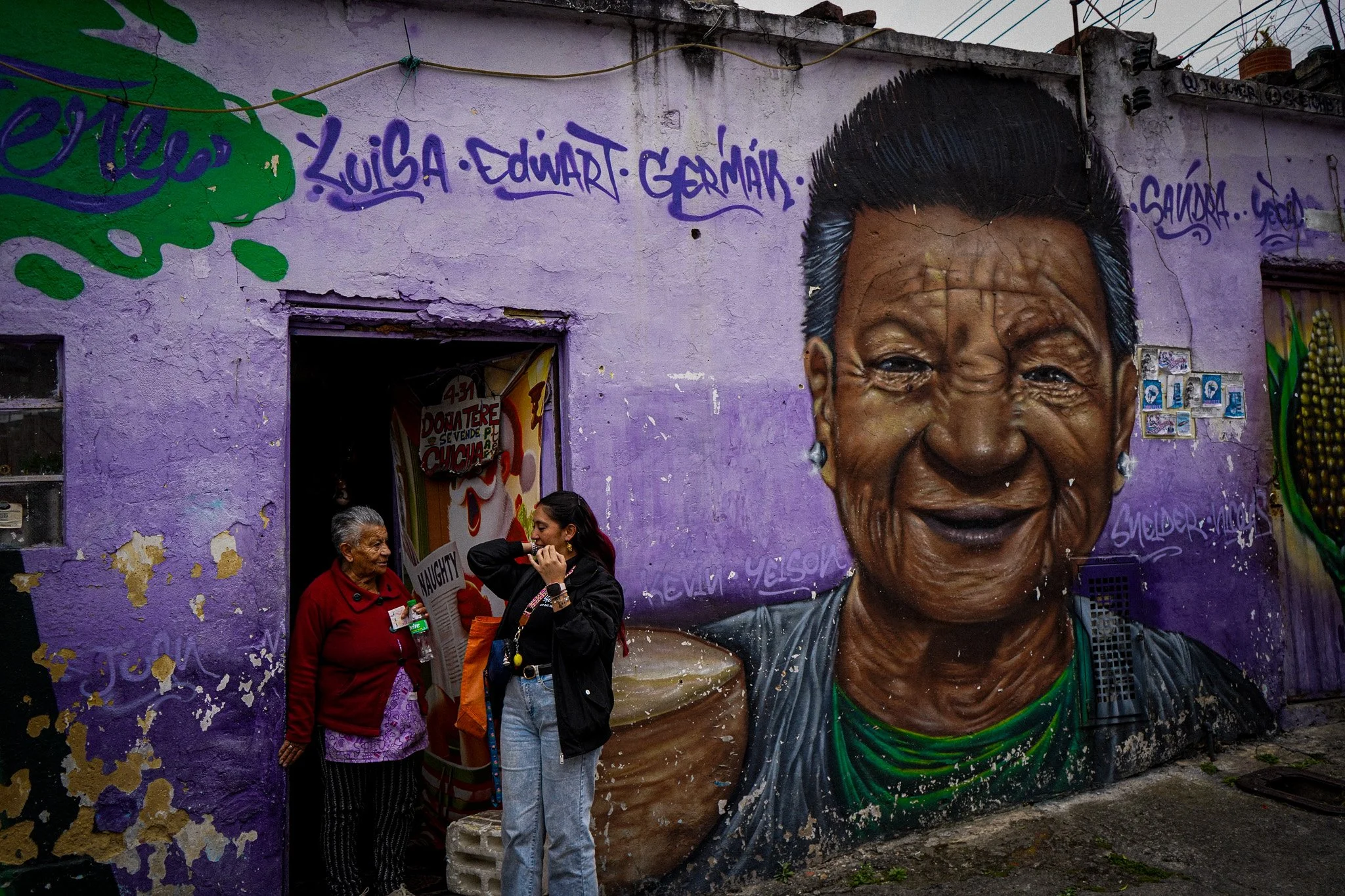Street scene with people standing in front of a wall with a large mural of an elderly woman's face and graffiti. Two women are talking, one older and one younger, near a jewelry stand and graffiti with names.