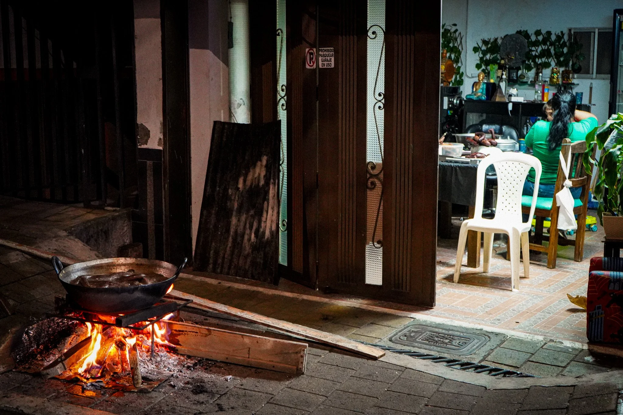 A cooking scene with an open fire and a wok, with a kitchen or outdoor cooking area visible in the background, including a person in a green shirt sitting at a table.