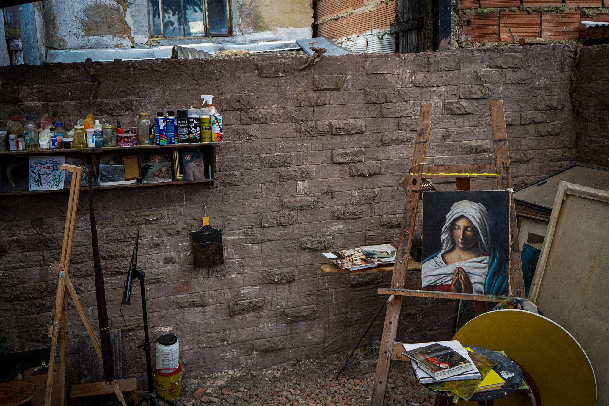 An outdoor art studio with a brick wall and a shelf holding various art supplies, including paints, brushes, and framed pictures. An easel displays a painting of a woman with a white headscarf. There is a table with art books, a yellow container, and