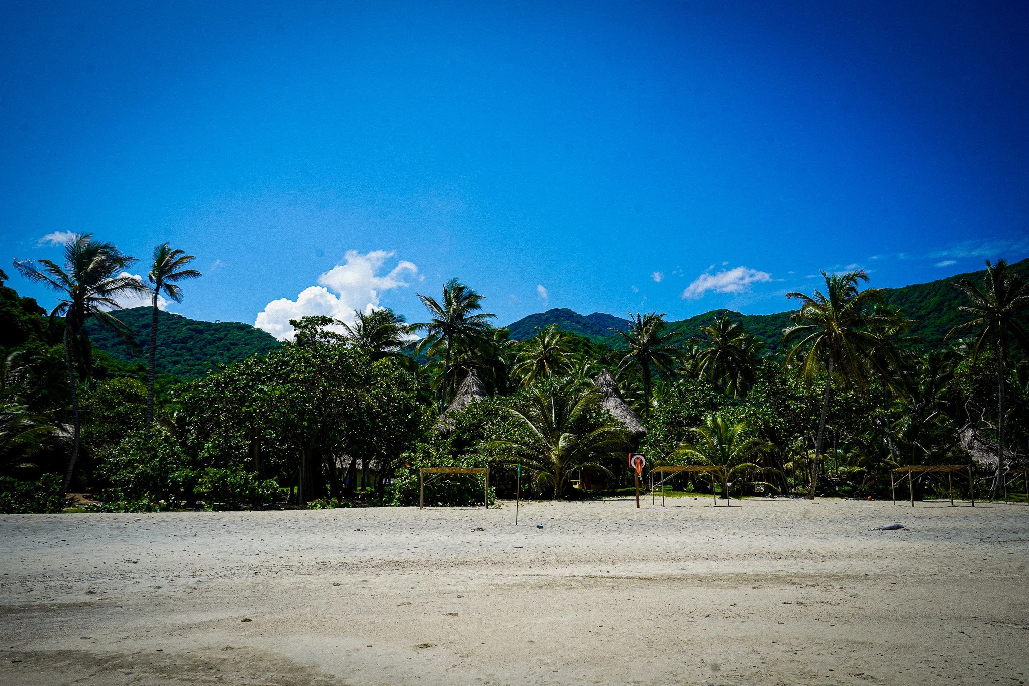 A beach with white sand, surrounded by lush green palm trees and mountains under a bright blue sky with a few clouds.