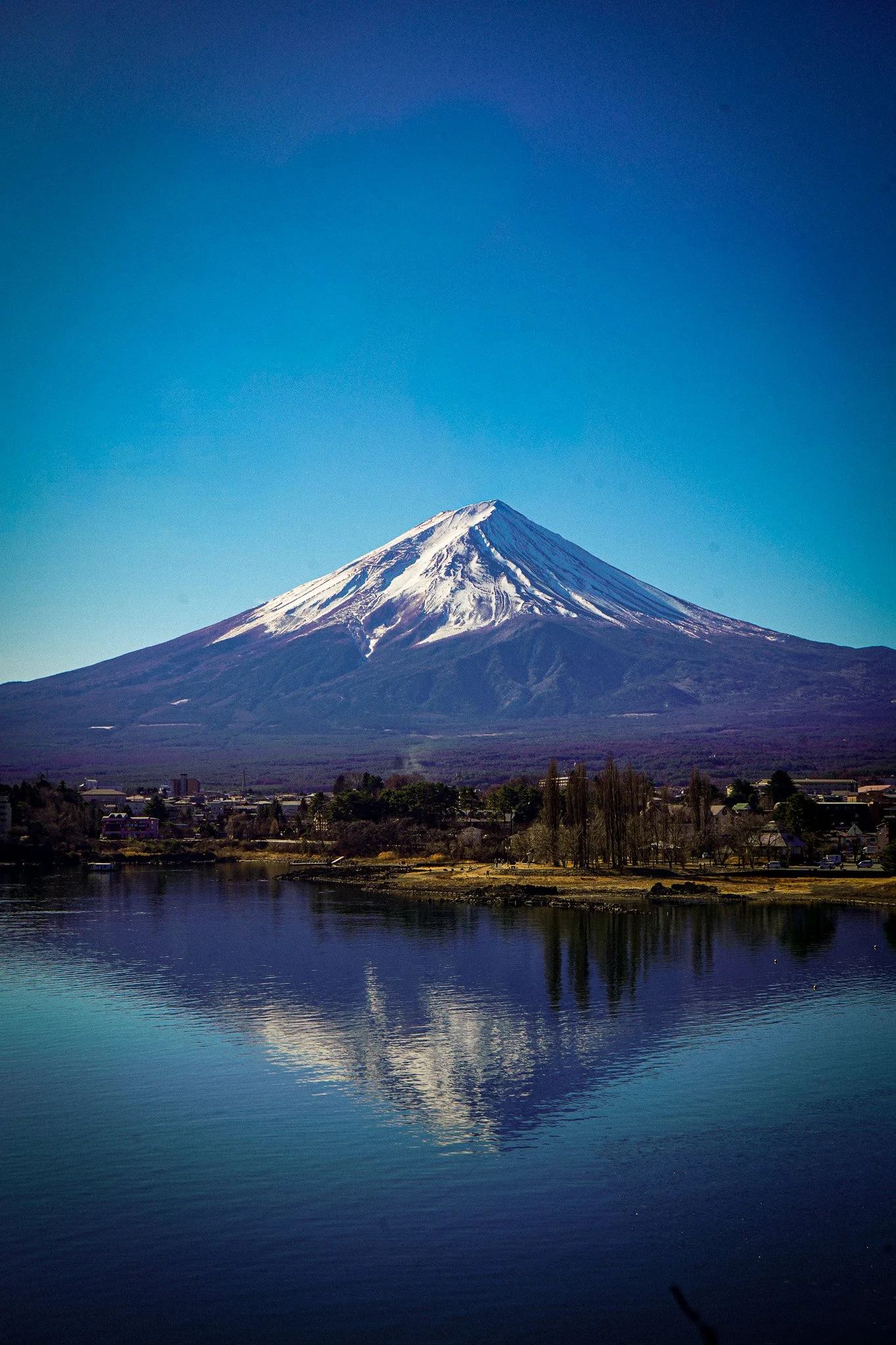 Snow-capped Mount Fuji with a clear blue sky and its reflection in a calm lake in the foreground.