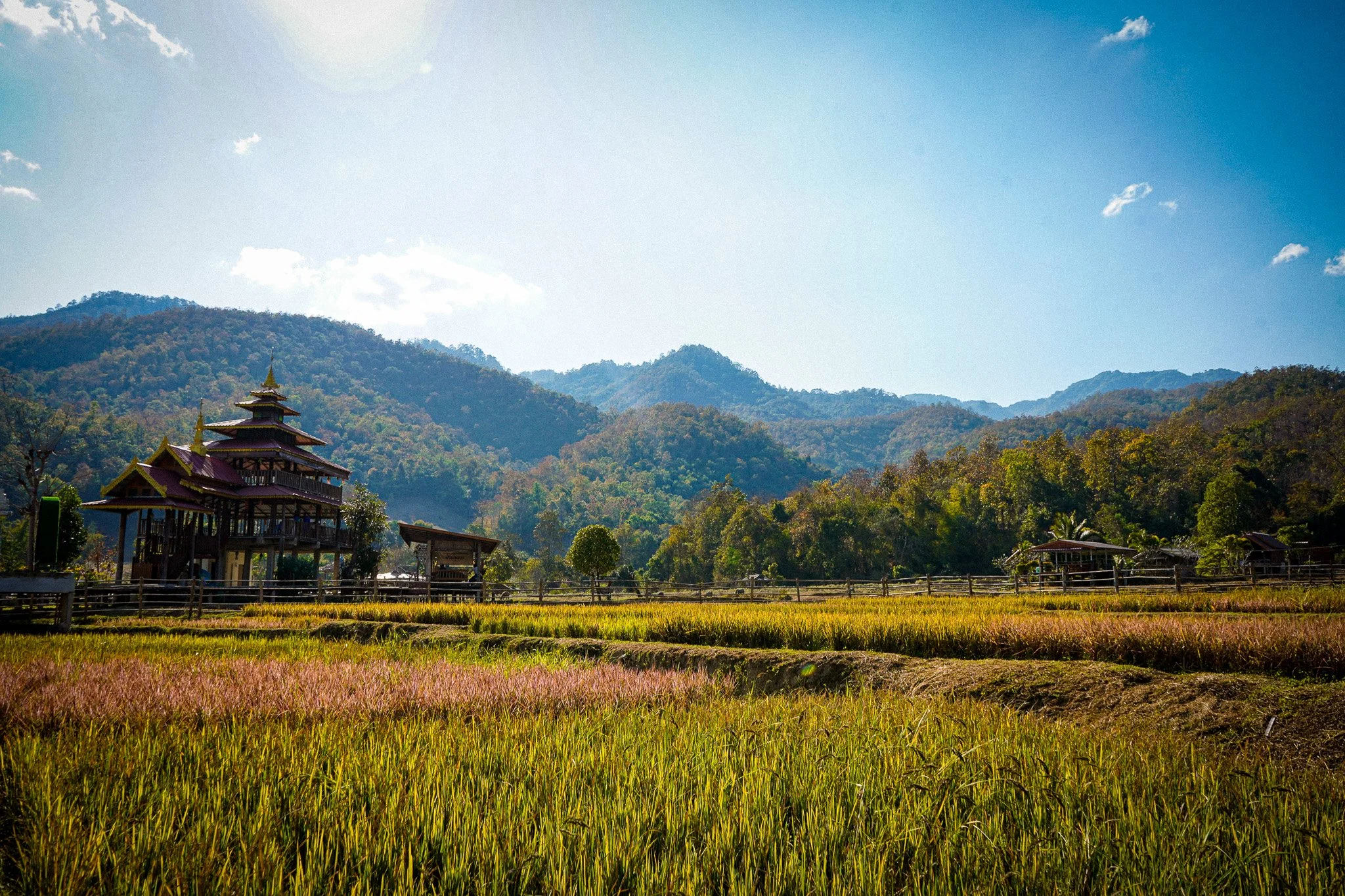 Scenic view of a mountainous landscape with a multi-tiered pagoda-style wooden structure in the foreground, surrounded by trees and fields under a partly cloudy blue sky.