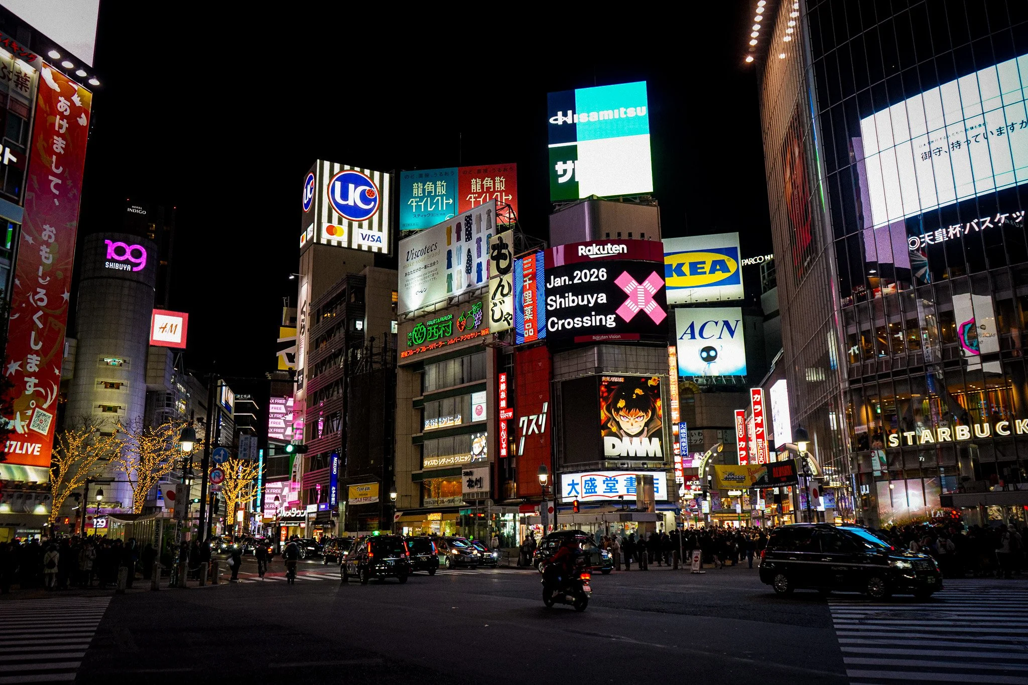 A busy city intersection at night with illuminated billboards and neon signs in Japanese and English, crowded with pedestrians and moving cars.