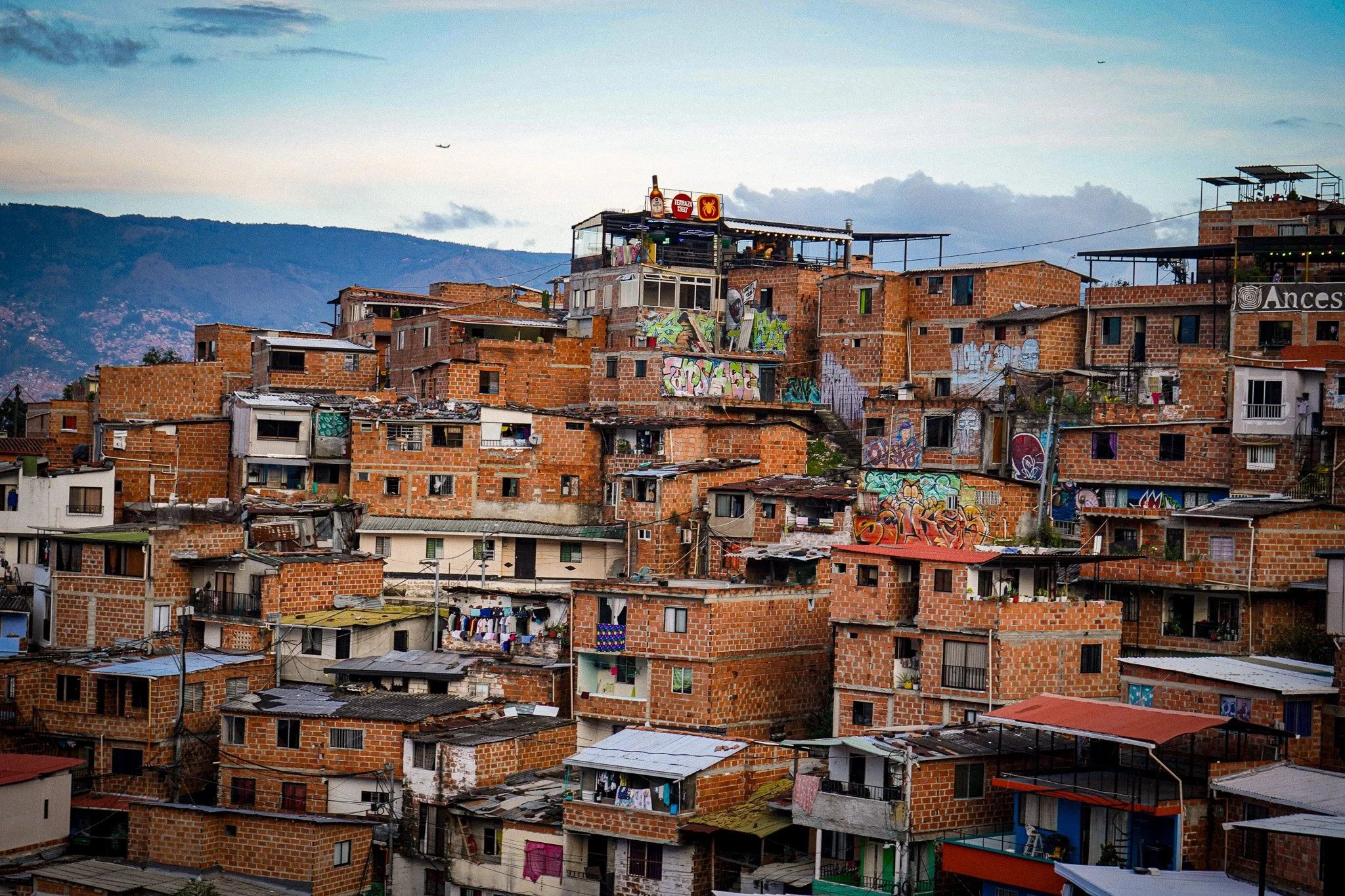 View of a hillside neighborhood with brick houses, some decorated with graffiti, under a sky with scattered clouds and airplanes flying overhead.