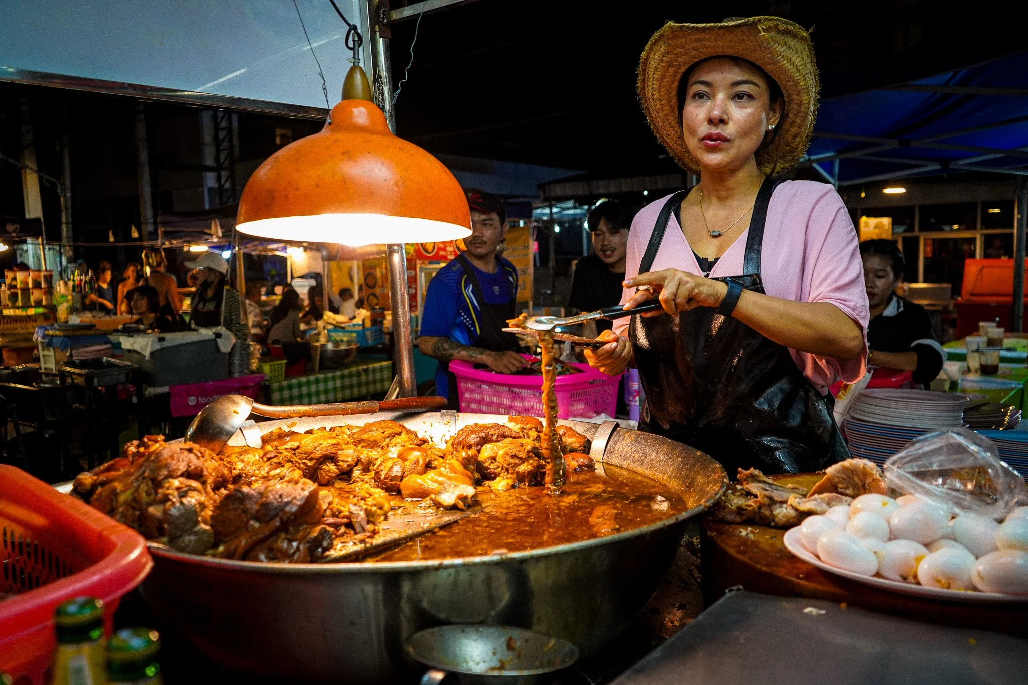 A woman wearing a straw hat and pink shirt serving food at a night market, with bowls of hard-boiled eggs and cooked meat in front of her.