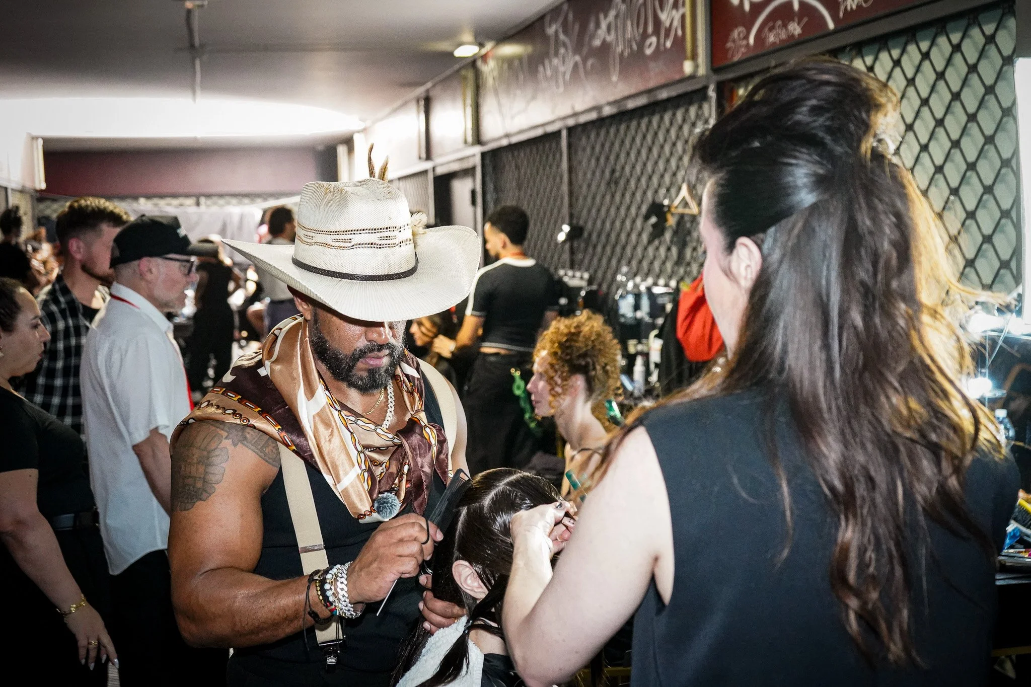 A man dressed in a colorful, patterned outfit with a large, white hat decorated with feathers, tattooed arms, and jewelry engages with a woman at a busy indoor event or convention. Several other people are visible in the background.