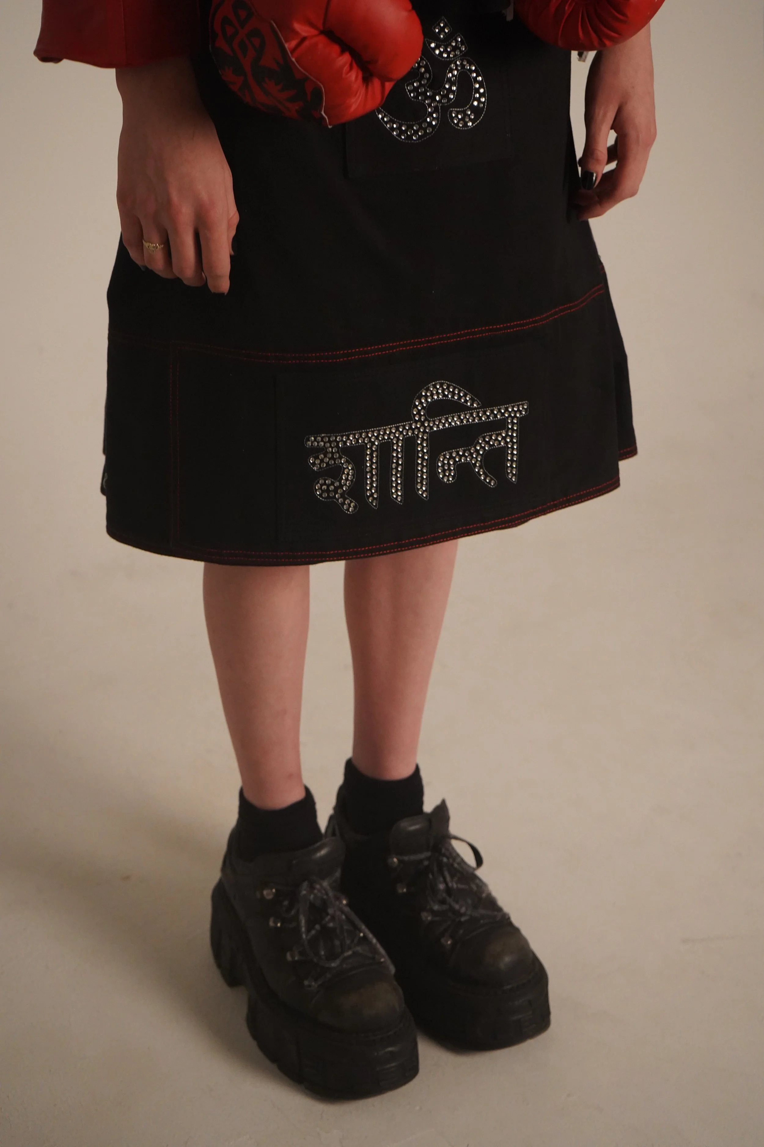 Person wearing black platform shoes, black socks, black skirt with red stitching and rhinestone embellishments, and a red jacket with black and red gloves.