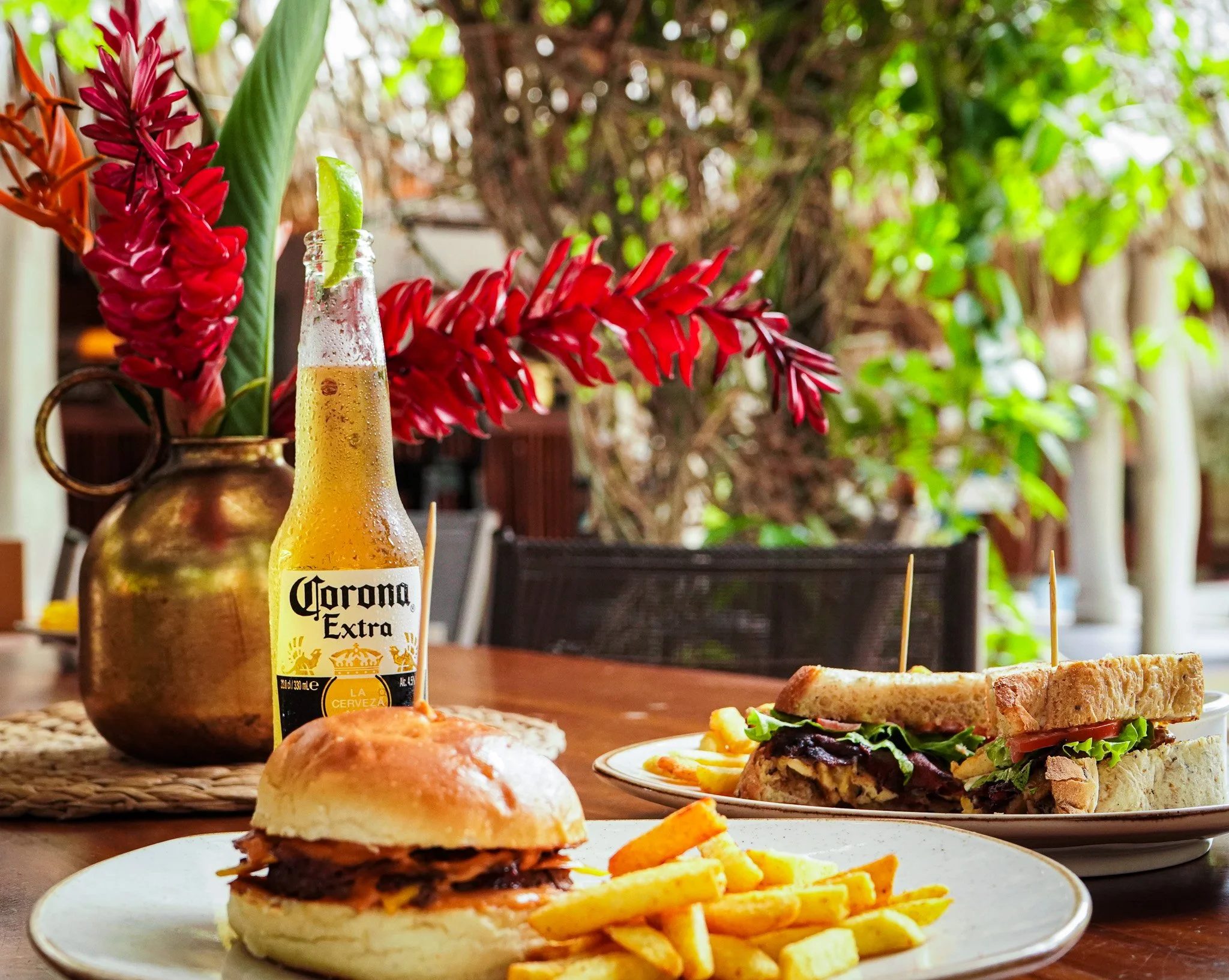 A table set with food and drinks, including a Corona beer bottle with lime, a burger with fries, a sandwich with lettuce, tomato, and turkey, and a small plate of fries, with a background of greenery and plants.