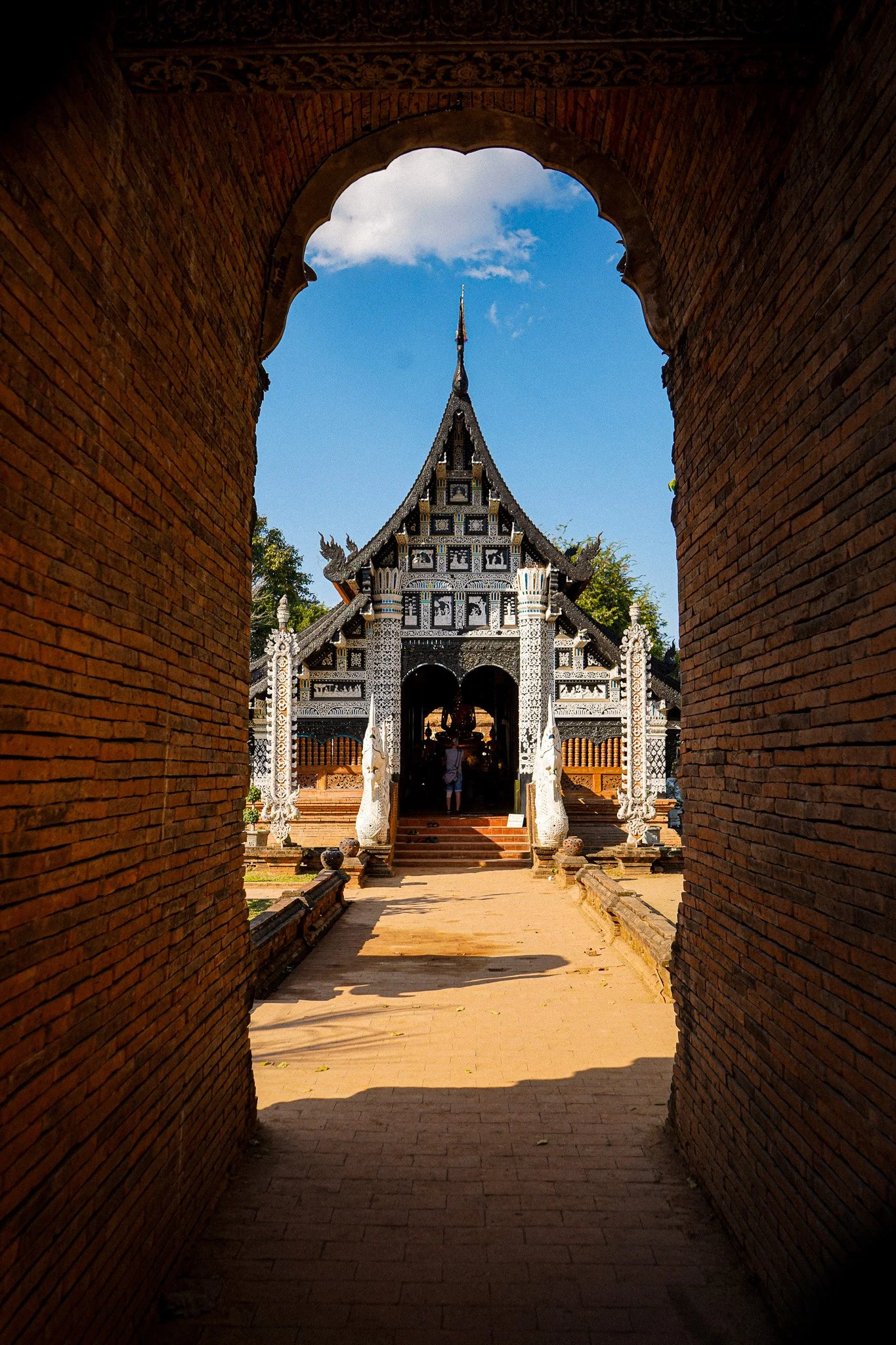 View through a brick archway showing a traditional Thai temple with ornate architecture under a blue sky with clouds.