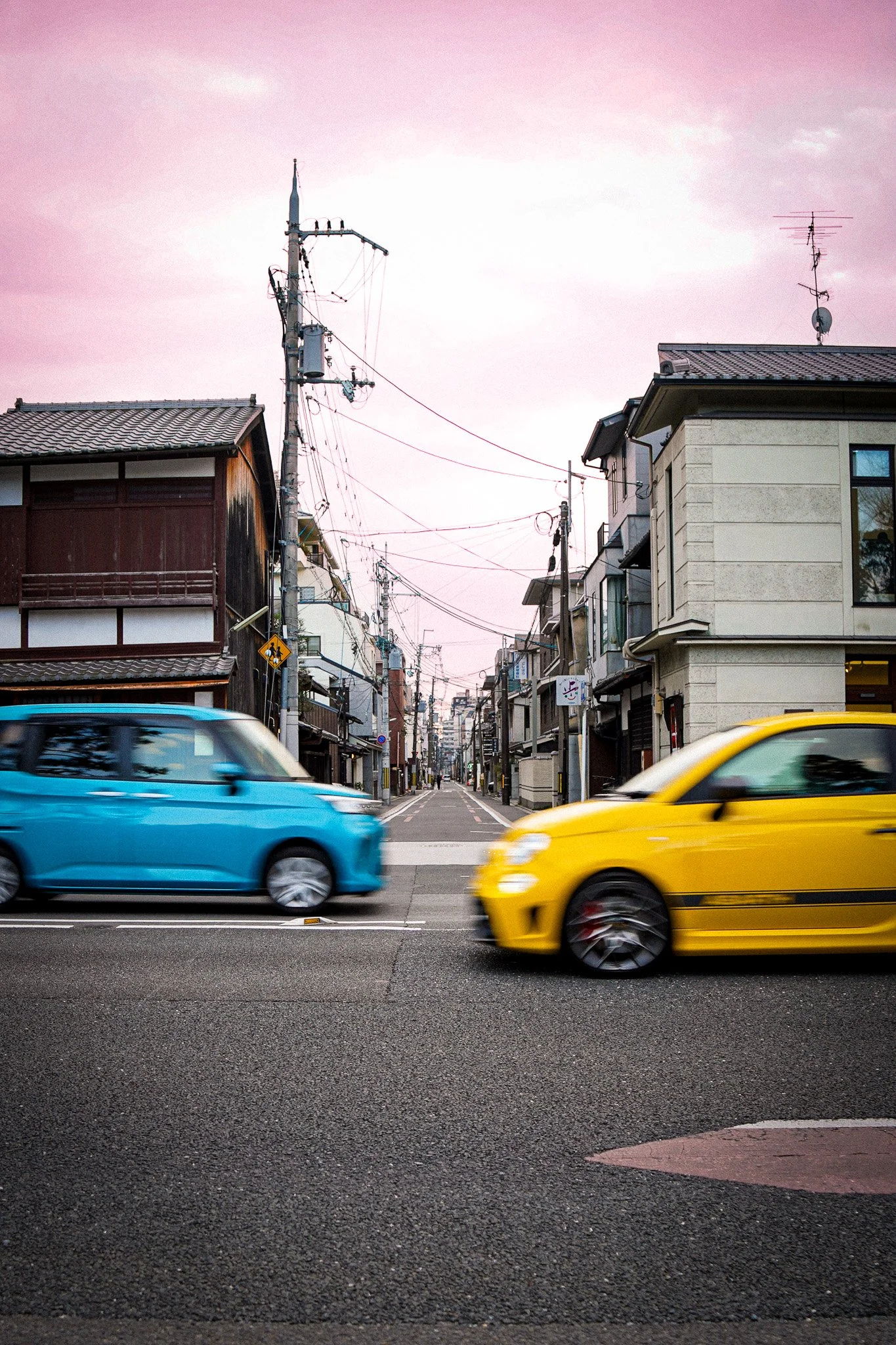 Two cars, one blue and one yellow, are moving side by side on a city street. There are buildings and power lines in the background, with a pink sky above.