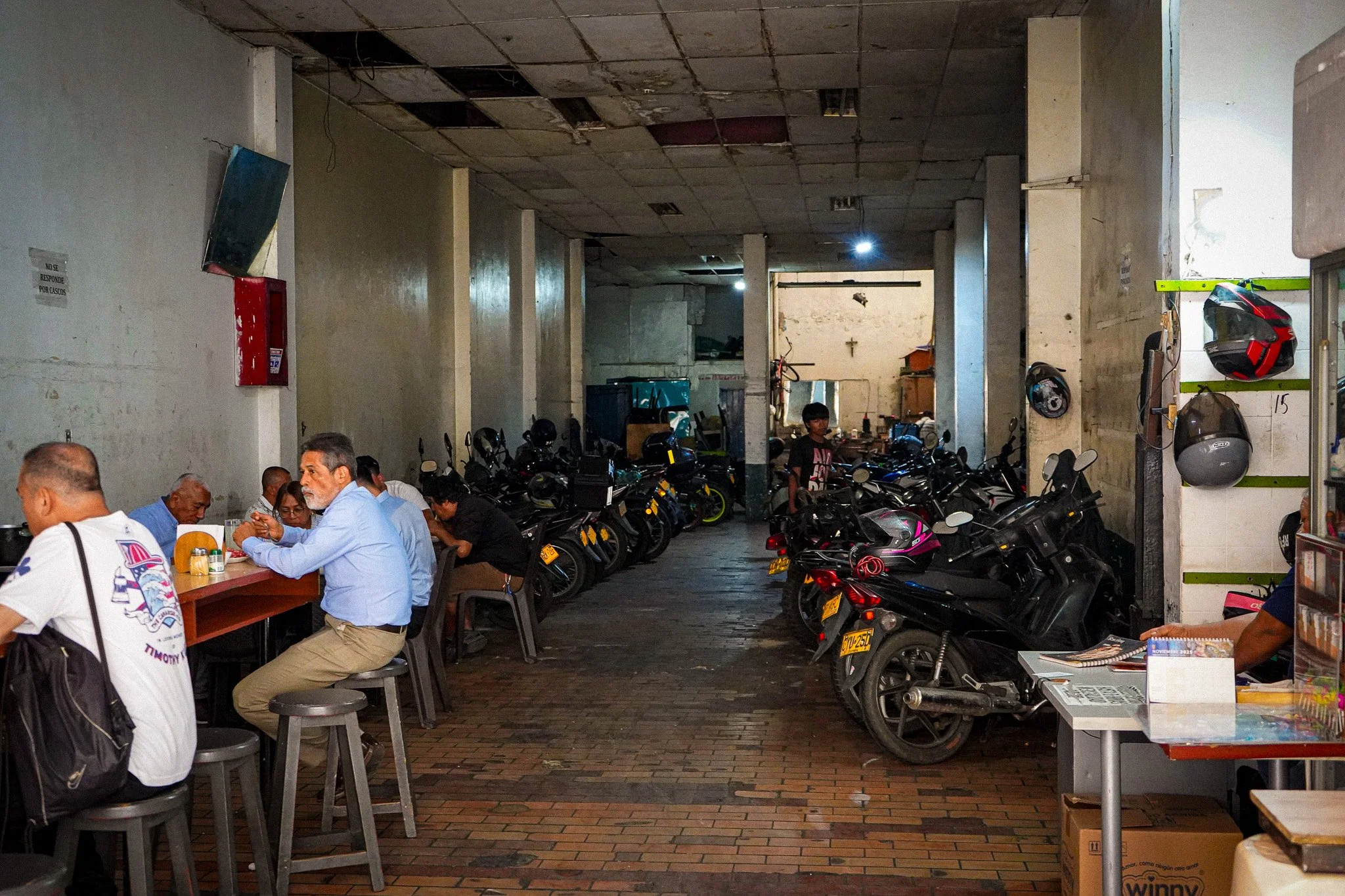 Indoor parking area with motorcycles and a few people sitting at tables, with unfinished ceiling and lighting