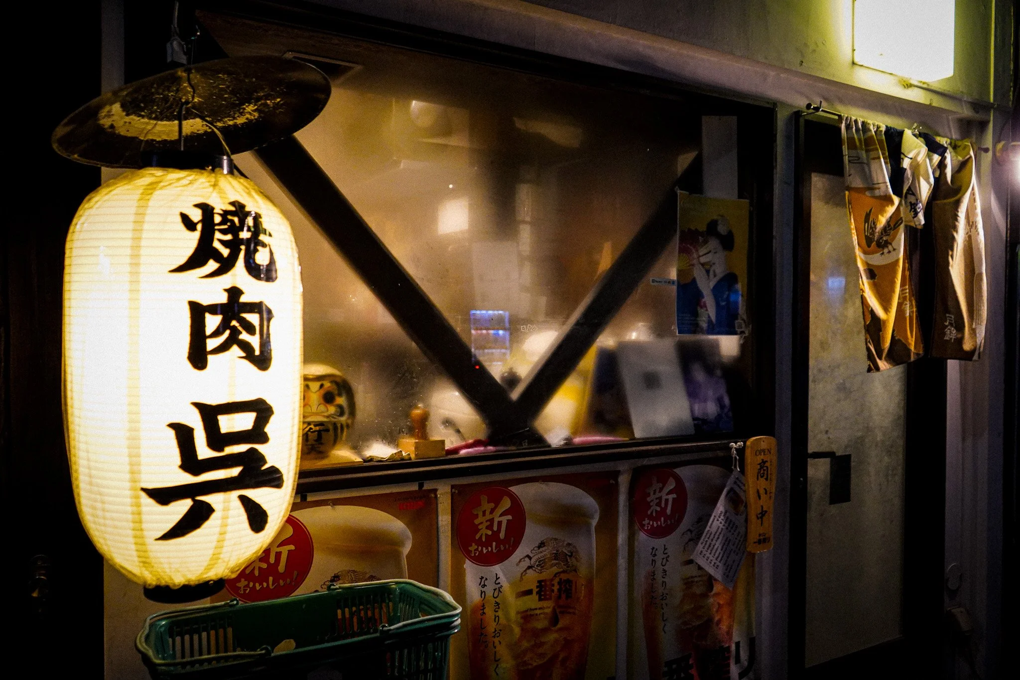 A Japanese paper lantern with black kanji characters hanging outside a restaurant, with a glass window displaying various menus and posters.