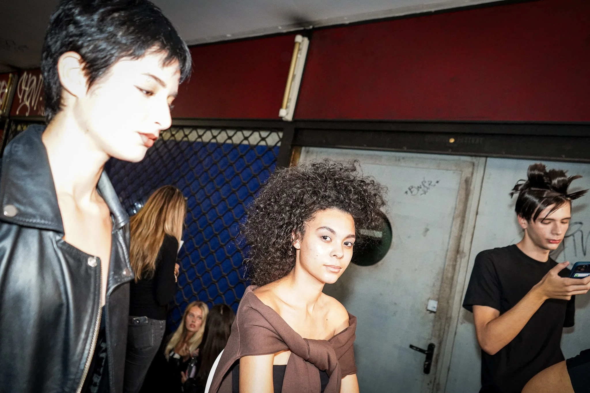 A group of young women standing together indoors. The woman in the foreground has dark, curly hair and is wearing a brown top. To her left, another woman with short black hair and a leather jacket is looking down. Behind them, another woman with ligh