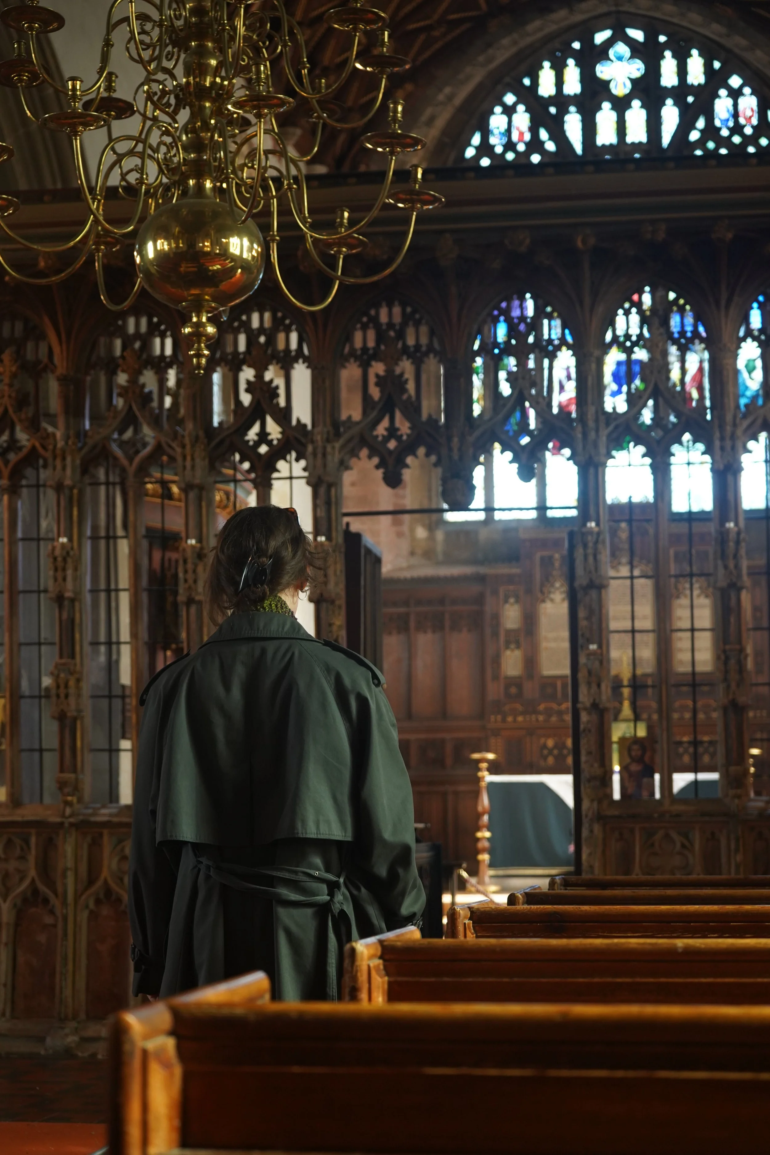 A person with brown hair wearing a black coat and headphones inside a church, facing away from the camera. The church has stained glass windows and an elaborate chandelier hanging from the ceiling.