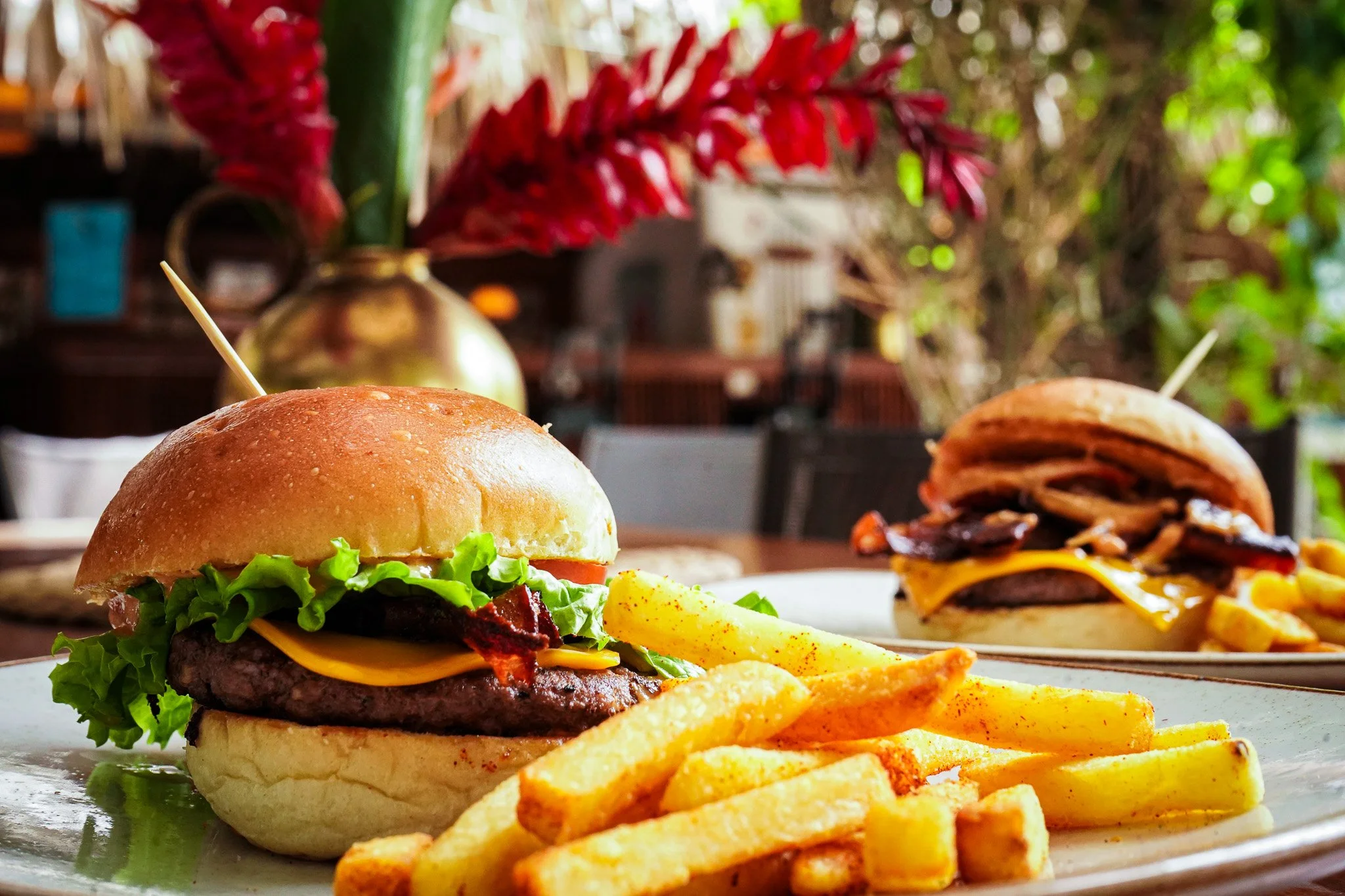 Close-up of two cheeseburgers with French fries on a white plate, with blurred background of a dining area and plants.