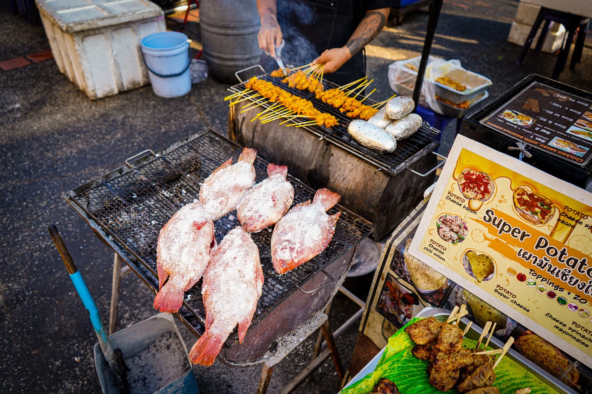 Street food stall with skewered marinated chicken grilling on a charcoal barbecue, whole fish with salt crust on a grill, and a menu display with various snack options.