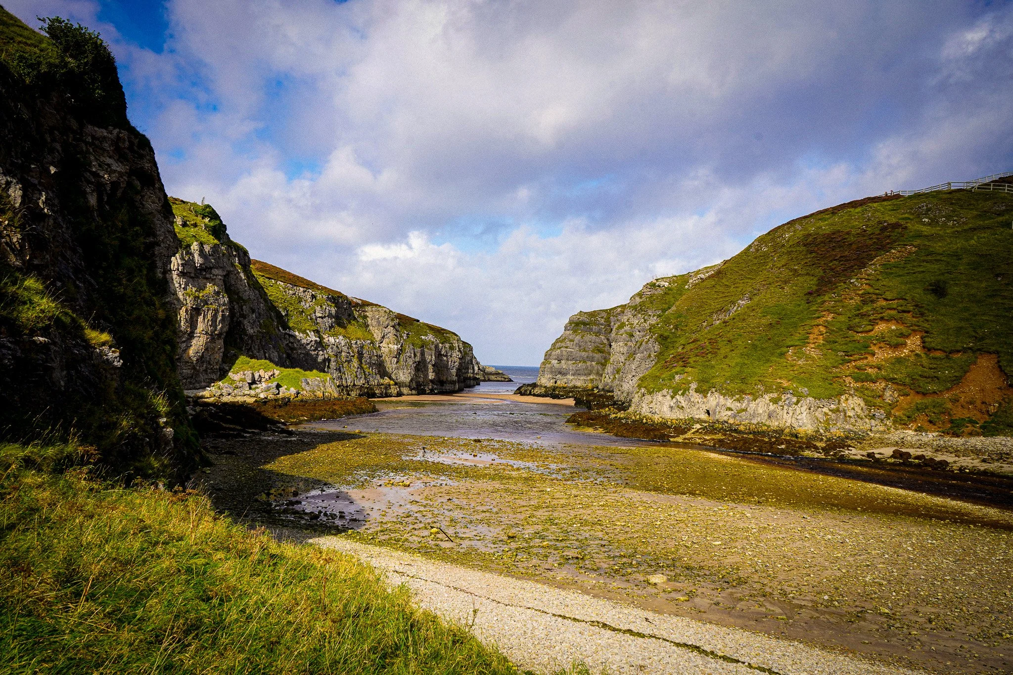 A coastal landscape with steep green hills, rocky cliffs, a narrow strip of sandy beach, and a calm body of water between the hills under a partly cloudy sky.