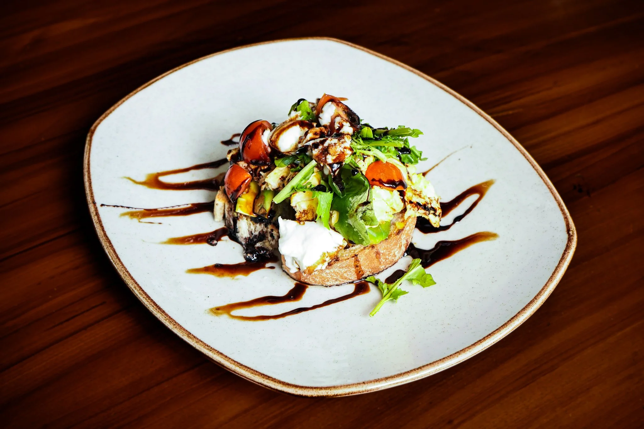 A plated dish with a piece of bread topped with salad greens, cherry tomatoes, herbs, cheese, and drizzled with balsamic glaze, on a white plate on a wooden table.