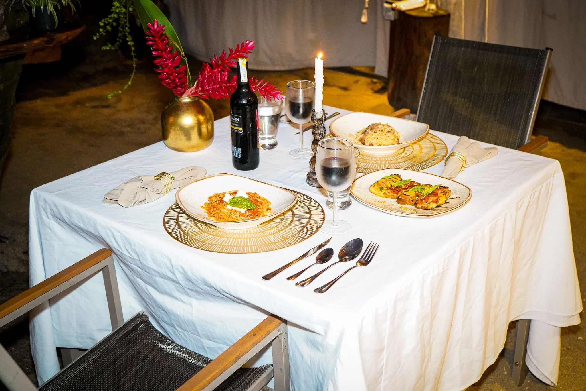 A table set for a meal with plates of pasta, glasses of water, a bottle of red wine, and a lit candle. There is a decorative gold vase with red flowers, a beige napkin with a gold napkin ring, and silverware arranged on a white tablecloth.