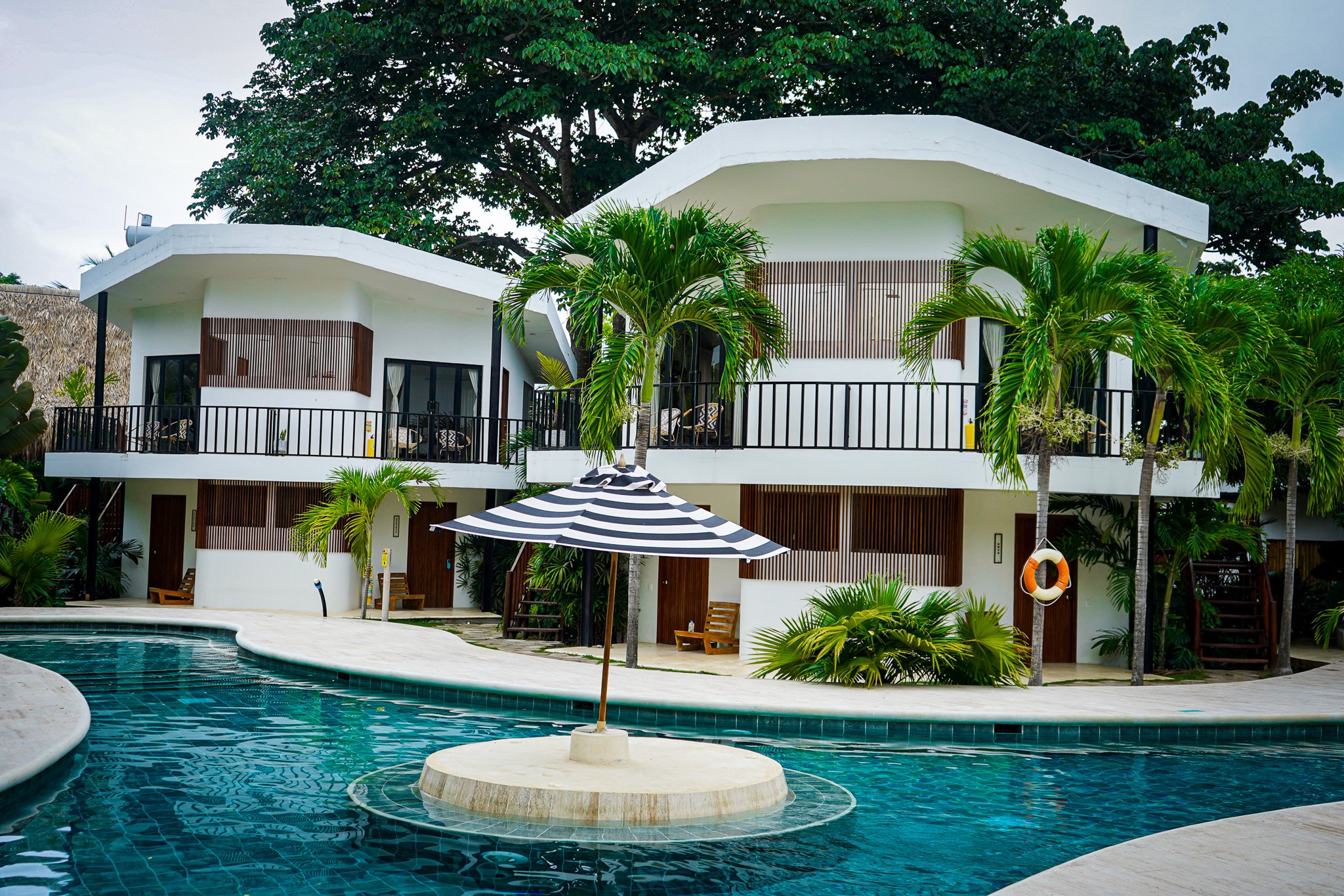 A modern white multi-story building with balconies surrounded by lush green palm trees and tropical plants, situated next to a swimming pool with a striped umbrella in the foreground.