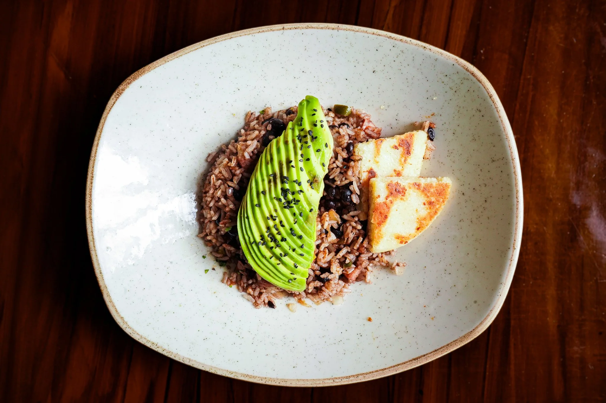A bowl of rice topped with slices of avocado and garnished with black sesame seeds, served with small pieces of grilled cheese on the side, on a wooden table.