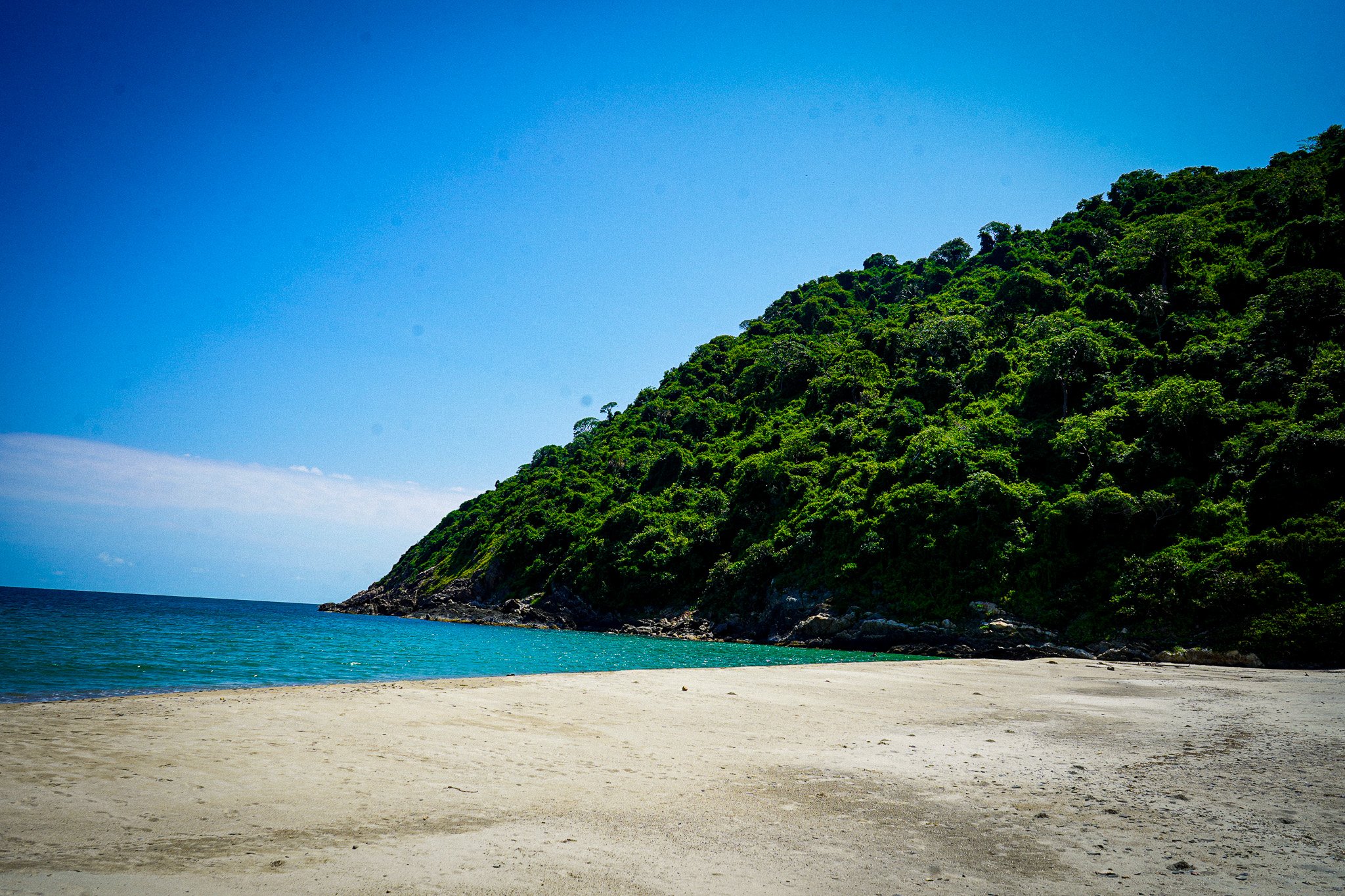 A scenic beach with a sandy shore, turquoise water, and a lush green hill covered with dense trees under a clear blue sky.