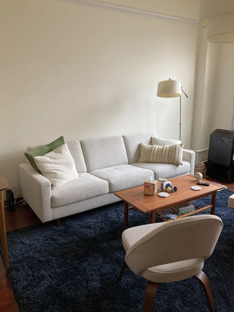 A photo of the therapy room that includes a cream coloured couch, a softly textured blue rug, an accent chair and coffee table with tissues and neuro affirming fidget toys.