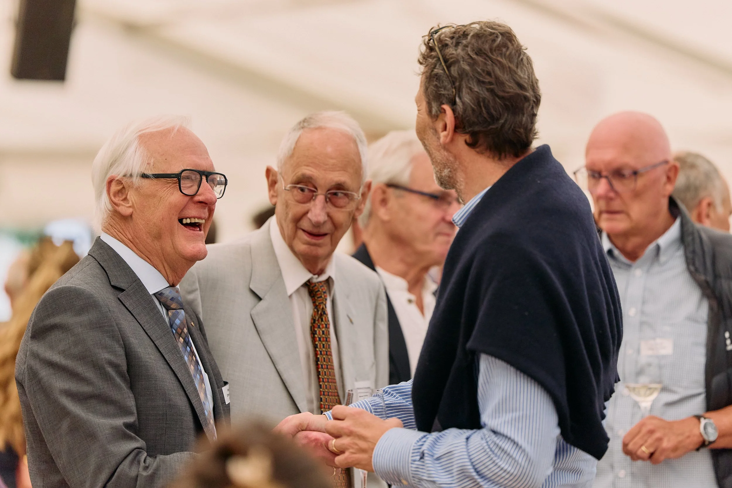 Mehrere ältere Männer im Gespräch, einer schüttelt einem jüngeren Mann die Hand bei einem formellen Treffen in einem Raum mit mehreren Teilnehmern. 100 Jahre Gymnasiums Friedberg Gossau SG.