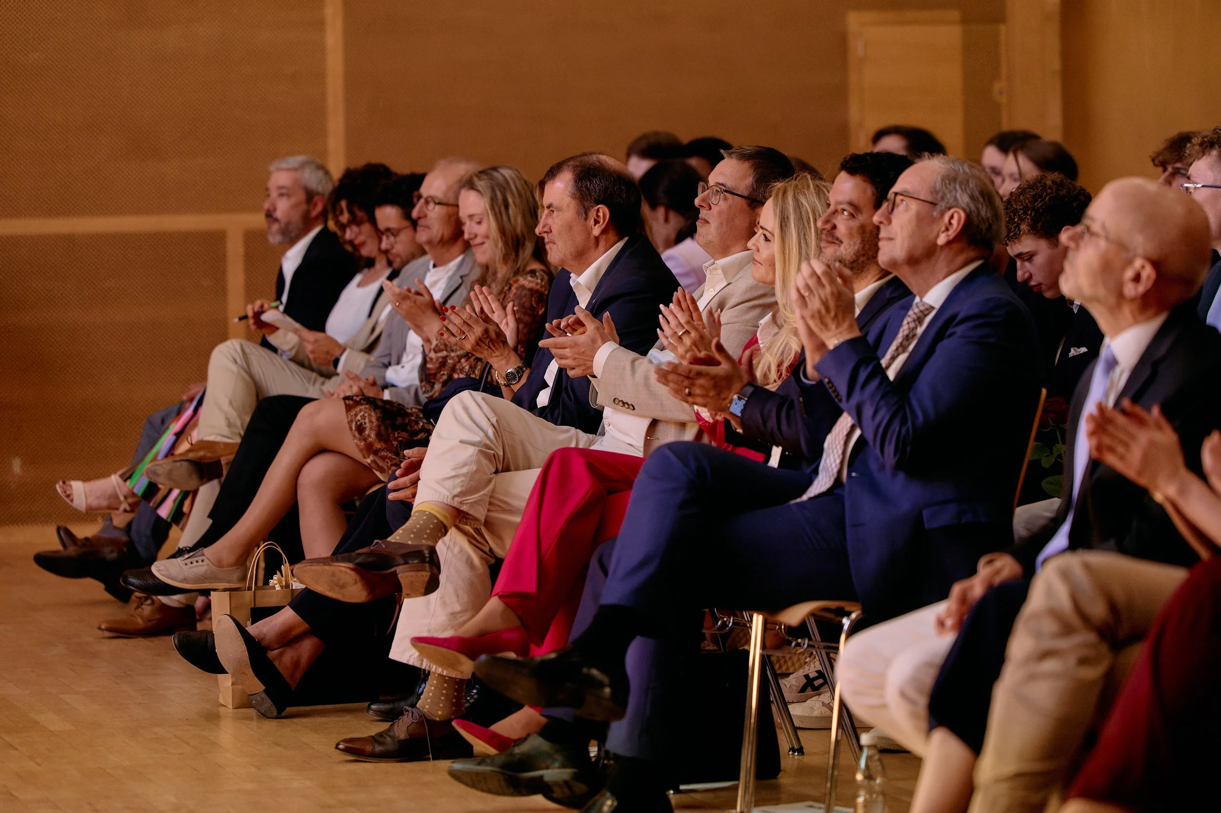 Zu sehen sind Menschen, die auf einem Konferenz- oder Seminar sitzen und sich offensichtlich eine Präsentation oder Rede ansehen, in einem Raum mit Holzverkleidung.