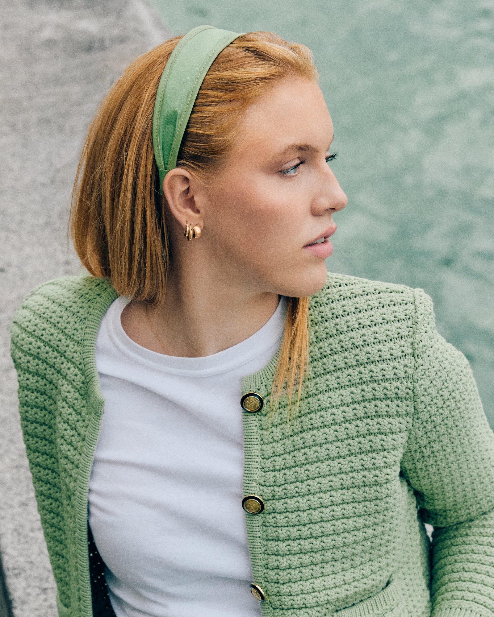 Nahaufnahme eines Mädchens mit roten Haaren, grüner Stirnband und Goldohrringen, trägt eine weiße T-Shirts und eine grüne Strickjacke, im Hintergrund Wasser und Steinflächen.