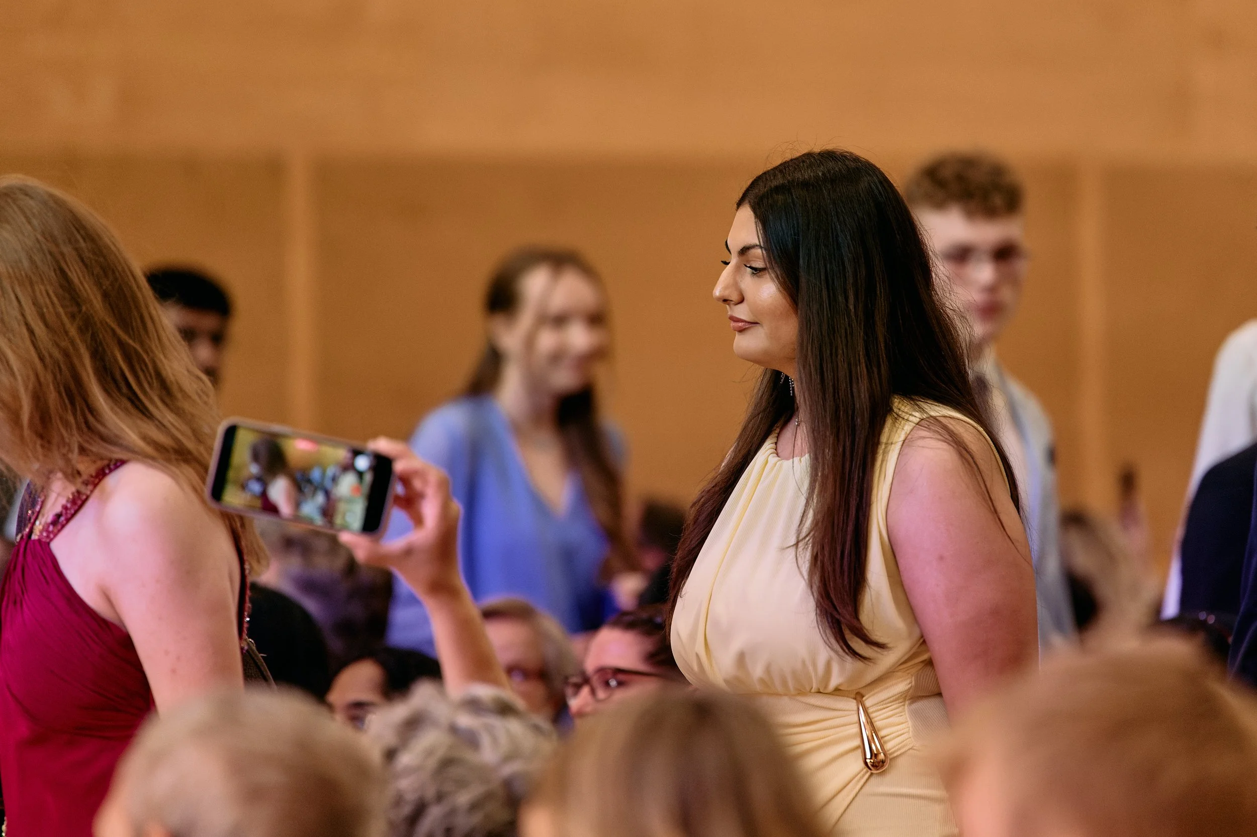 Zwei Frauen stehen sich gegenüber, eine macht ein Foto von der anderen mit einem Smartphone. Im Hintergrund sind weitere Personen bei einer Veranstaltung.