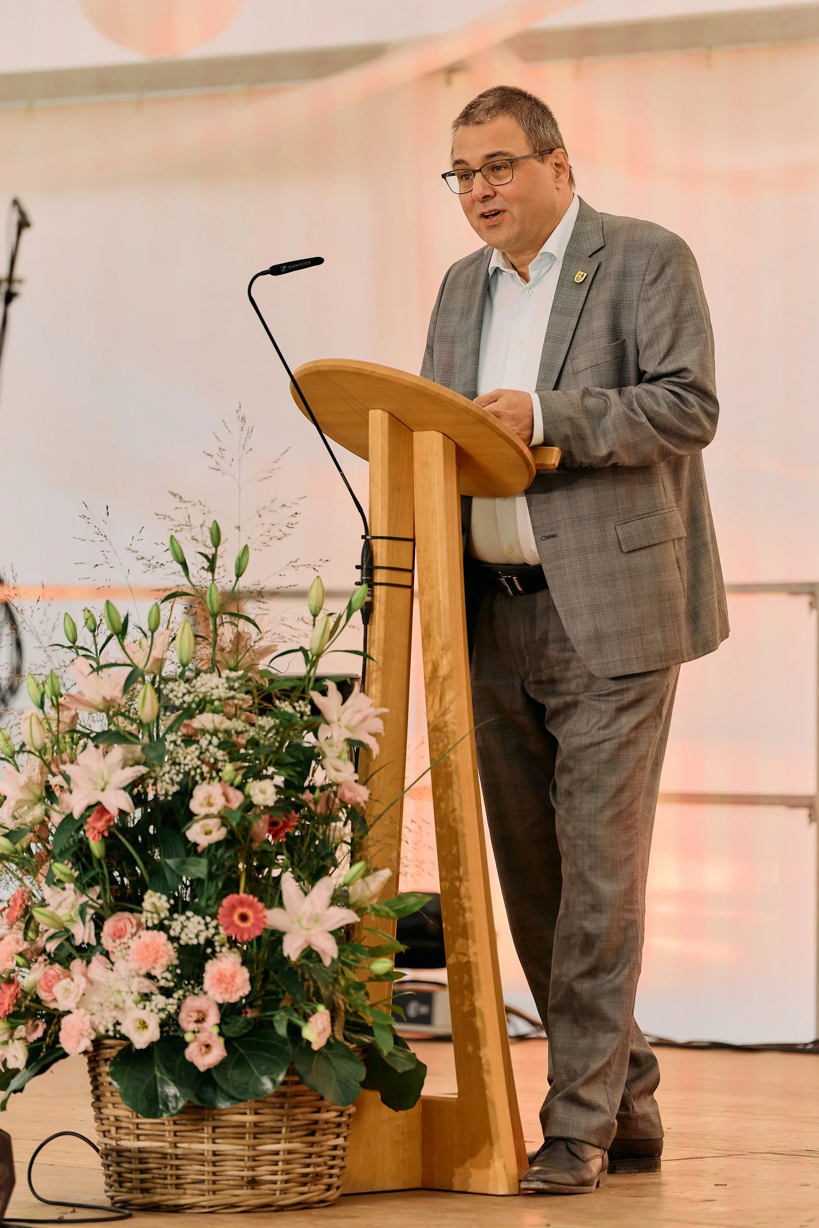 Ein Mann in einem grauen Anzug spricht an einem Rednerpult bei einer Veranstaltung, im Vordergrund befindet sich ein großer Blumenstrauß in einem Korb. 100 Jahre Gymnasiums Friedberg Gossau SG. Stadpräsident Gossau Wolfgang Giella