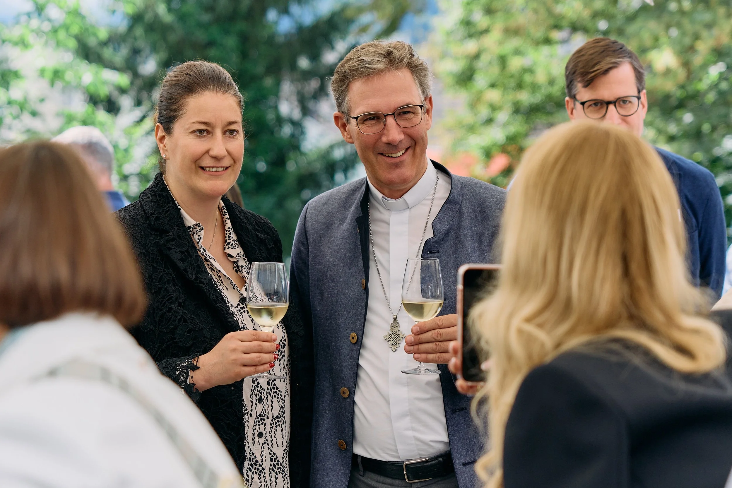 Fotograf & Videograf Wil SG | Businessfotografie Ostschweiz Daniel Dorrer Drei Menschen bei einer Feier im Freien, die mit Wein in der Hand lächeln, im Hintergrund Bäume und Natur. 100 Jahre Gymnasiums Friedberg Gossau SG.
