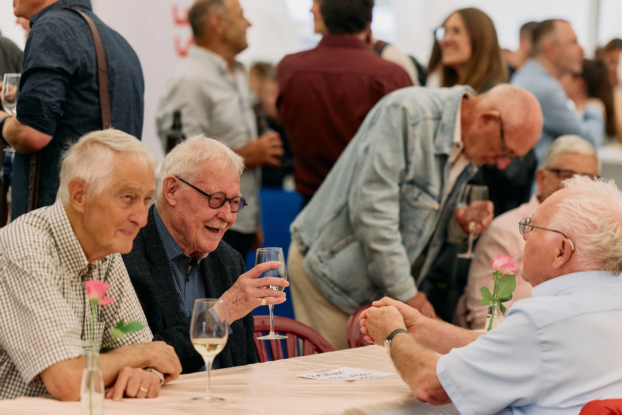 Menschen bei einer sozialen Veranstaltung, es findet eine Begrüßung oder Händeschütteln statt, im Vordergrund sind ältere Männer, einige trinken Wein, im Hintergrund unterhalten sich weitere Personen. 100 Jahre Gymnasiums Friedberg Gossau SG.