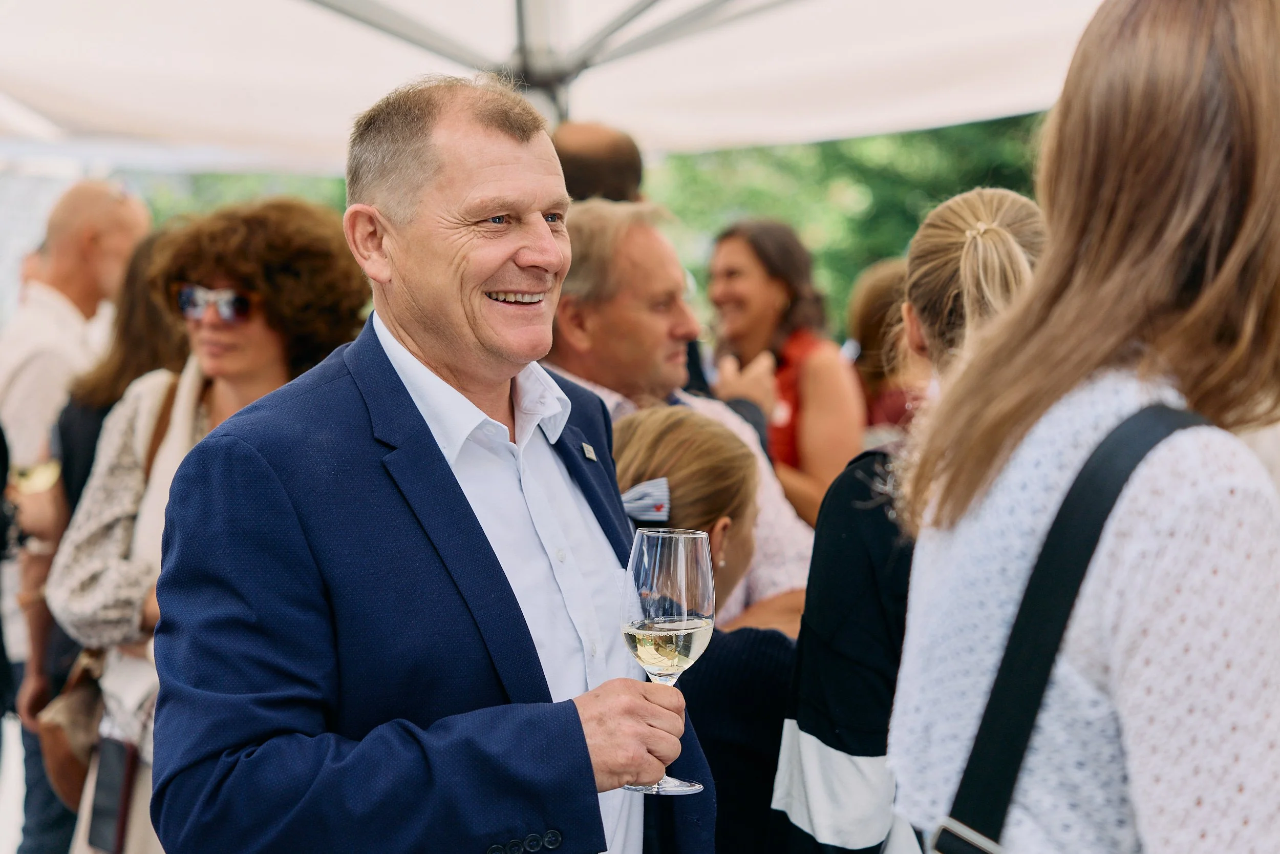 Fotograf & Videograf Wil SG | Businessfotografie Ostschweiz Daniel Dorrer Ein Mann in einem blauen Anzug hält ein Glas Sekt und lächelt auf einer Feier im Freien, umgeben von anderen Menschen. 100 Jahre Gymnasiums Friedberg Gossau SG.
