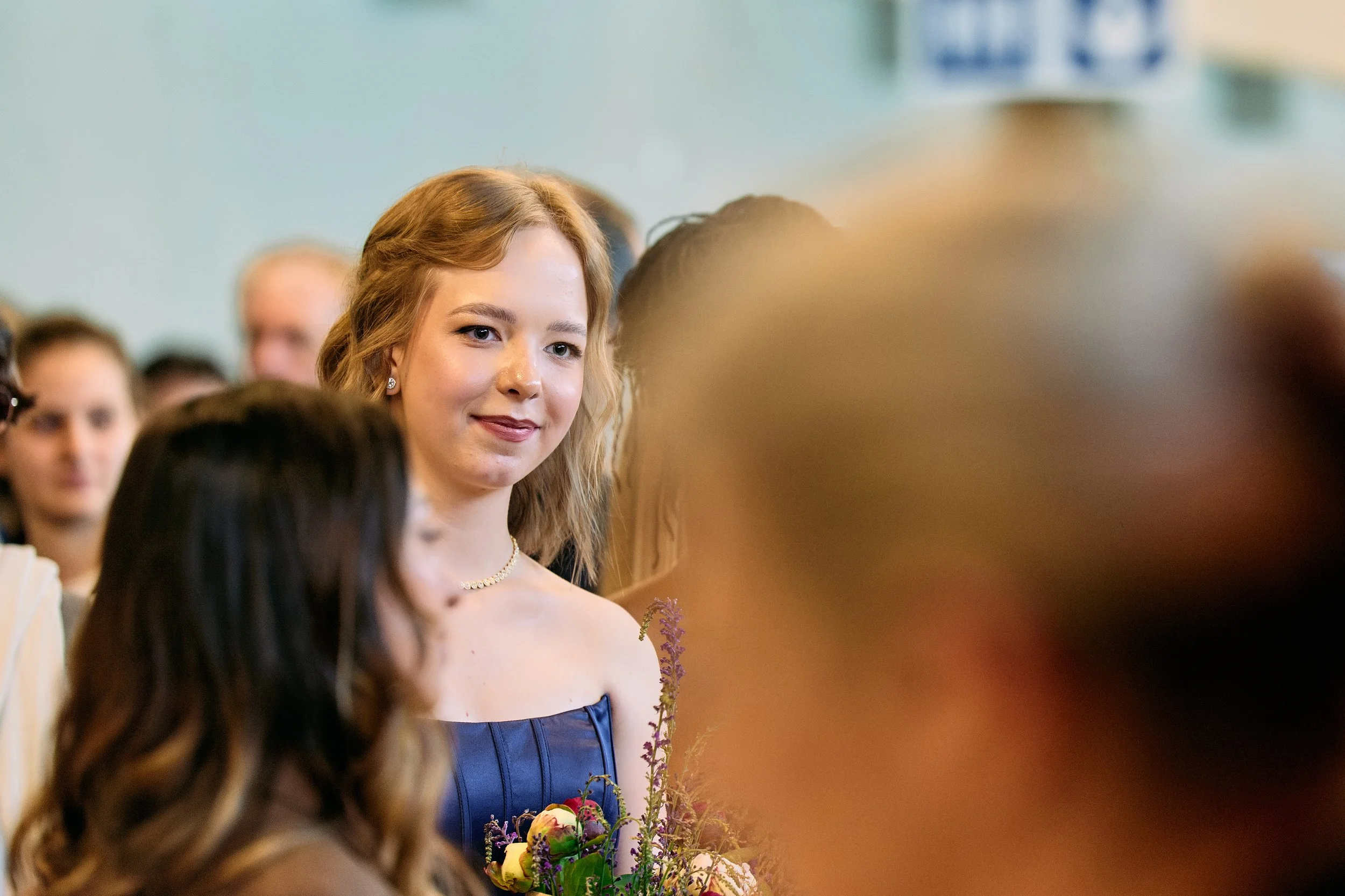Eine junge Frau mit lockigem, rotem Haar trägt eine elegante dunkle Abendrobe und ein Collier. Sie steht in einem Raum voller Menschen und hält einen Blumenstrauß. Die Szene wirkt festlich.