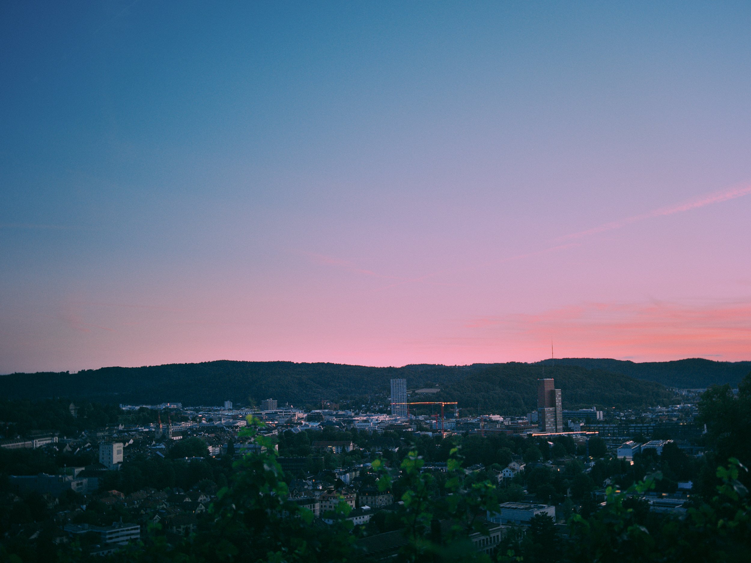 Stadtansicht bei Sonnenuntergang mit bläulichem Himmel, Bergen im Hintergrund und Verlauf der Stadt im Vordergrund.