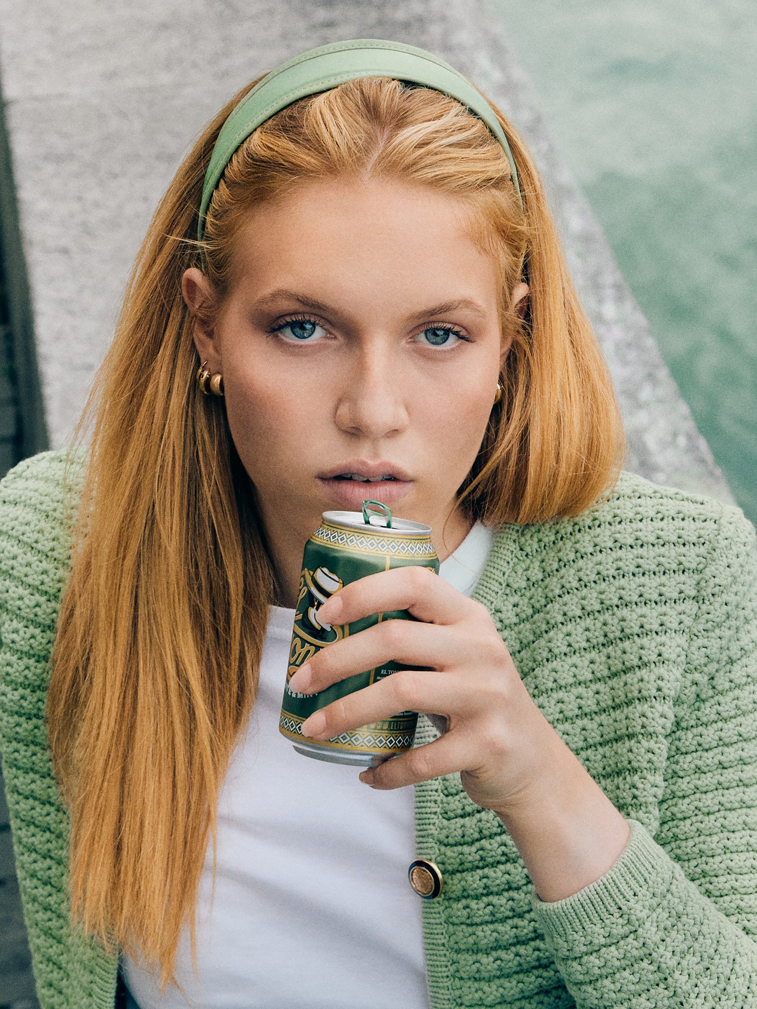 Eine junge Frau mit roten Haaren und blauen Augen hält eine Dose in der Hand, trägt eine grüne Strickjacke und einen weißen Pullover. Sie sitzt an einem Wasserweg und schaut direkt in die Kamera.