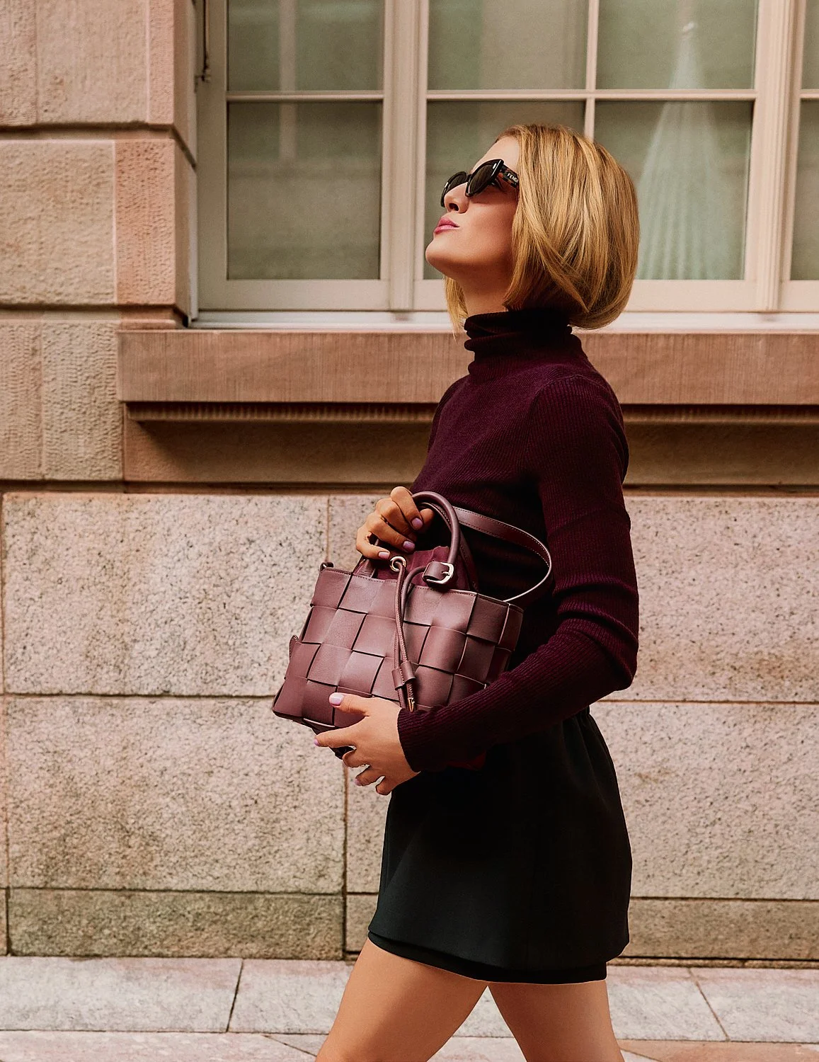 Eine stilvolle Frau mit kurzen, blonden Haaren trägt eine Sonnenbrille, einen dunkelroten Rollkragenpullover und einen schwarzen Rock. Sie hält eine braune Handtasche und läuft an einer Gebäudewand mit großen Fenstern vorbei.