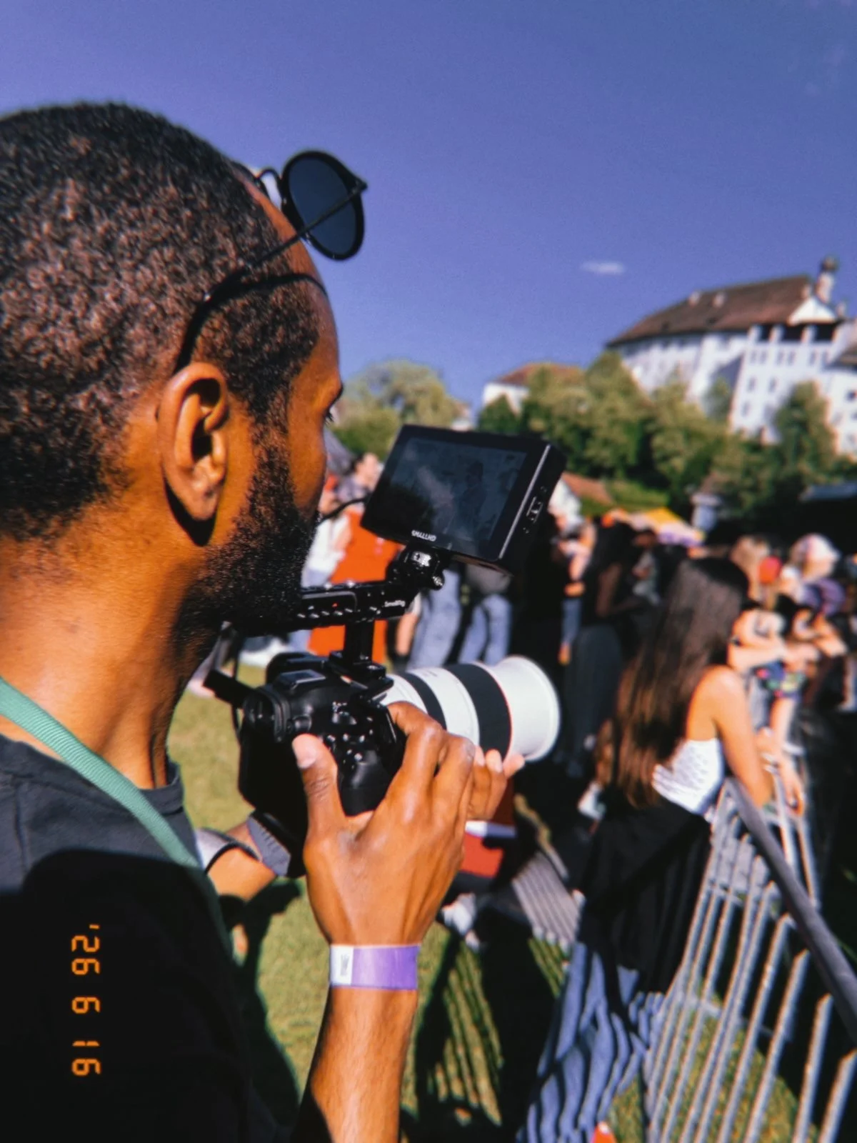 Fotograf mit Kamera bei einem Outdoor-Event, Menschen im Hintergrund, auf einem Hügel mit Gebäuden, blauer Himmel.