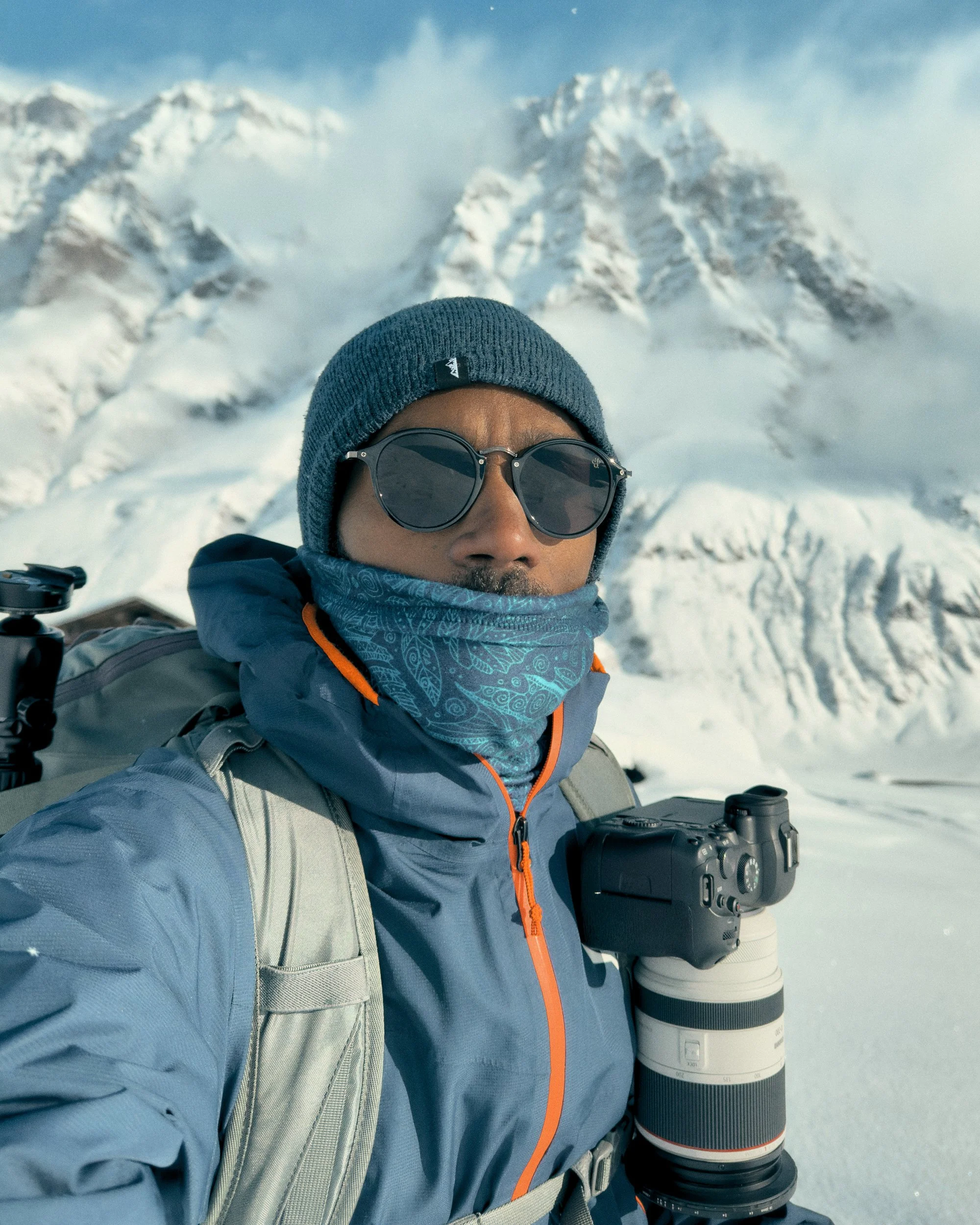 Mann trägt Winterkleidung, inklusive Mütze, Sonnenbrille, Halswärmer und Rucksack, vor schneebedeckten Bergen und Schnee.